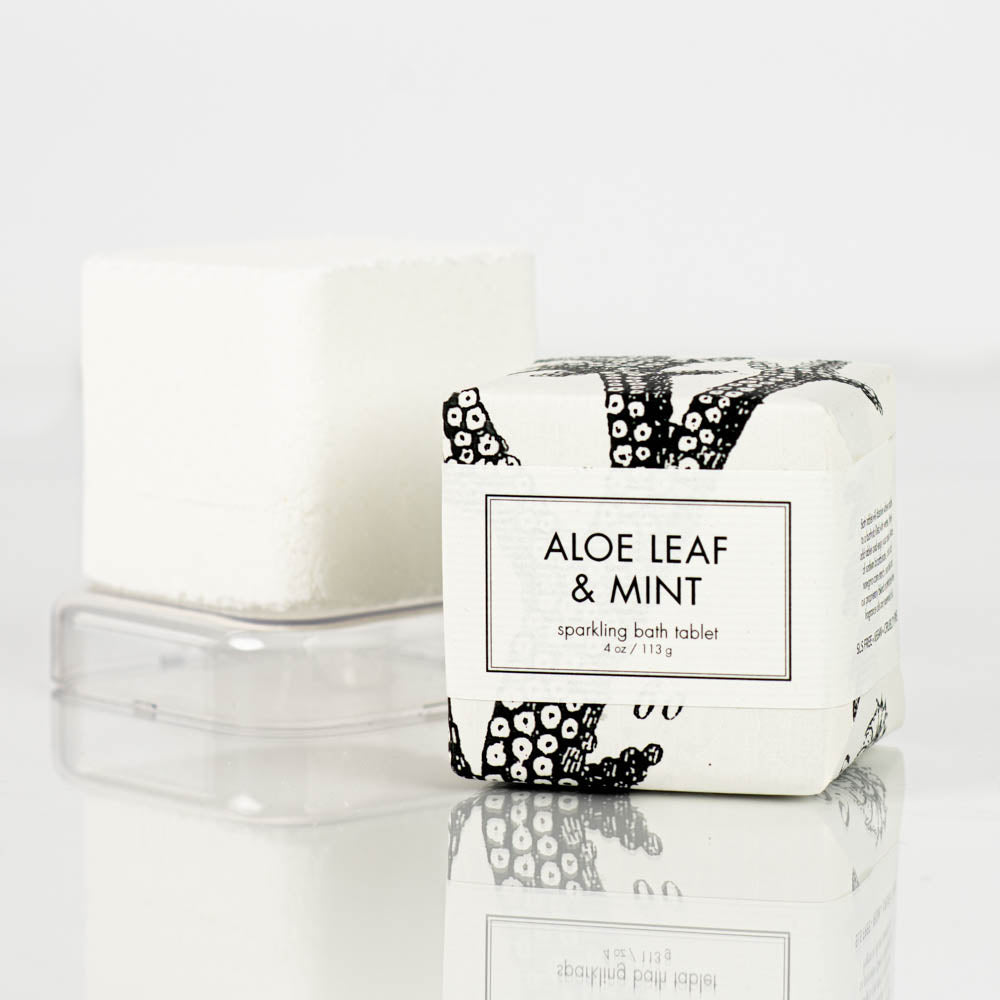 A Formulary55 Aloe Leaf and Mint Sparkling Bath Bomb Tablet, wrapped in white paper adorned with black designs, is placed beside an unwrapped white bath bomb on a clear plastic base. The packaging displays the text "Aloe Leaf & Mint".