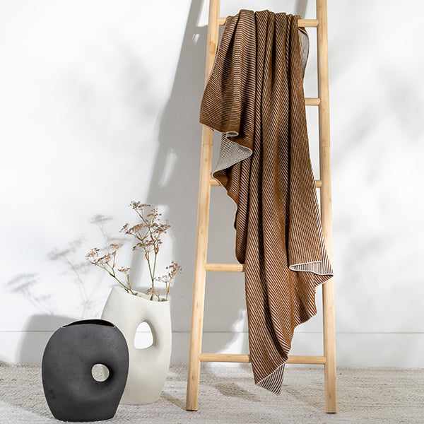Copper textured organic cotton blanket draped over a wooden ladder against a white wall with decorative vases.