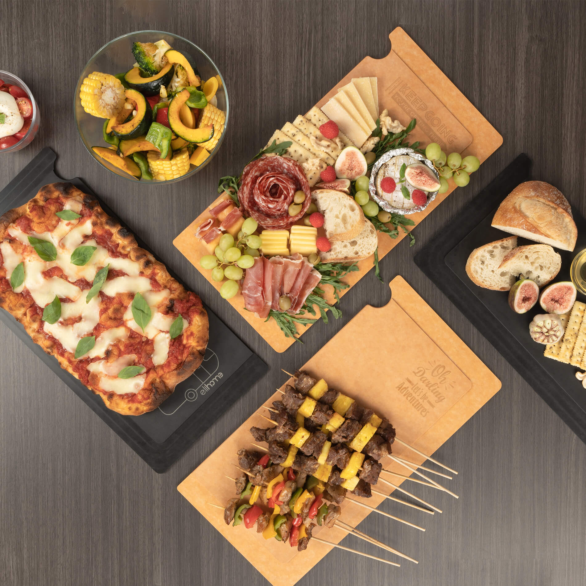 Overhead view of Elihome custom Airstream RV sink covers used as elegant serving platters. Features a black heat-resistant board with pizza and natural wood fiber boards holding charcuterie and shish kabobs, showcasing the "Oh Darling" and "Keep Going" engraved designs.