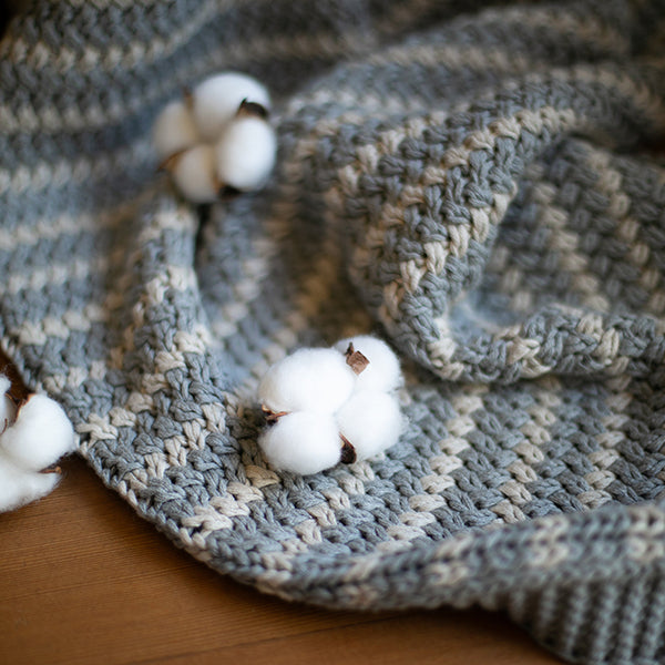 Light gray and white knit cotton throw blanket with cotton balls on a wooden surface.