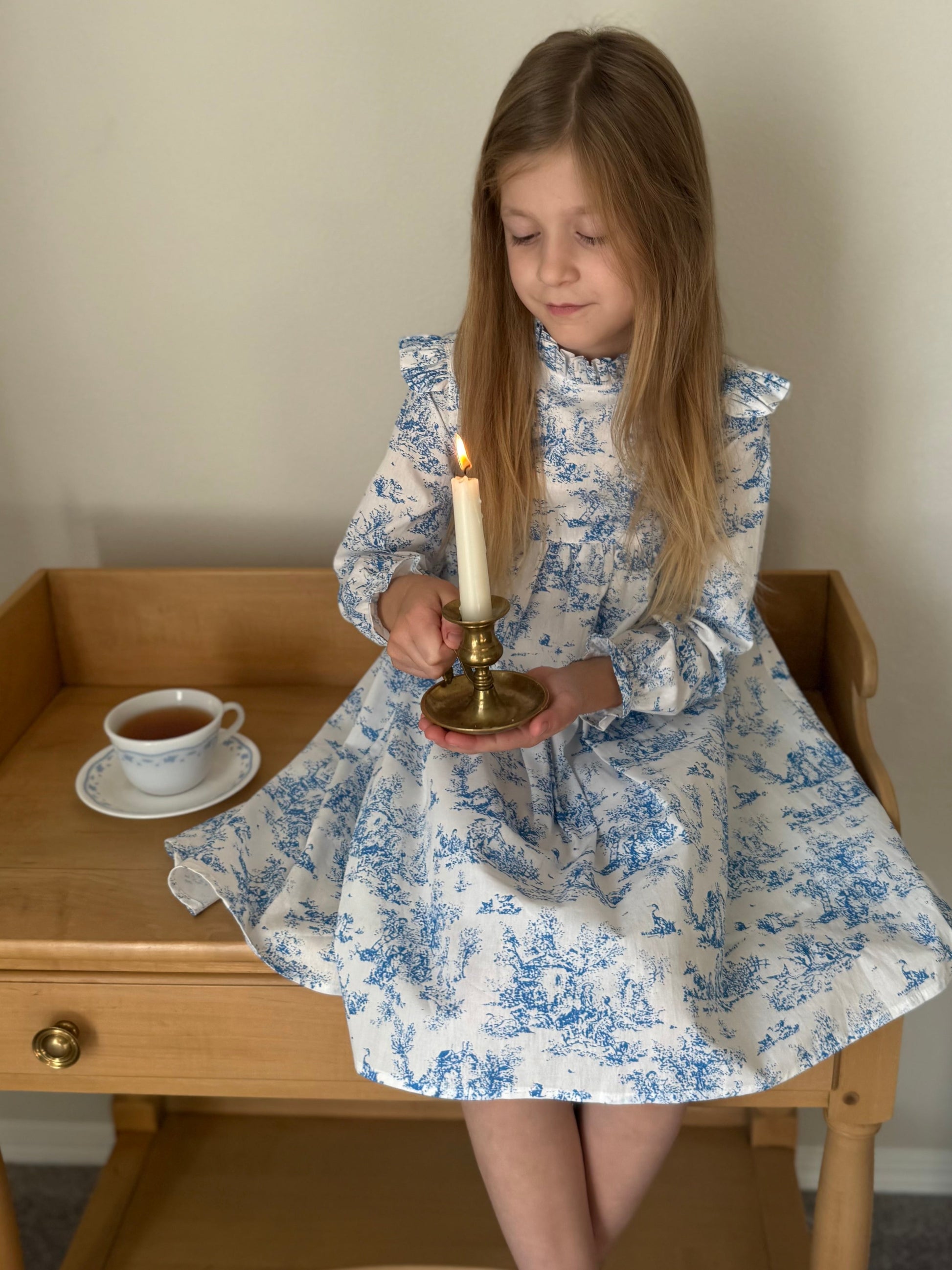 Young girl in a blue toile floral print  dress holding a candle, sitting on a wooden table with a cup of tea.