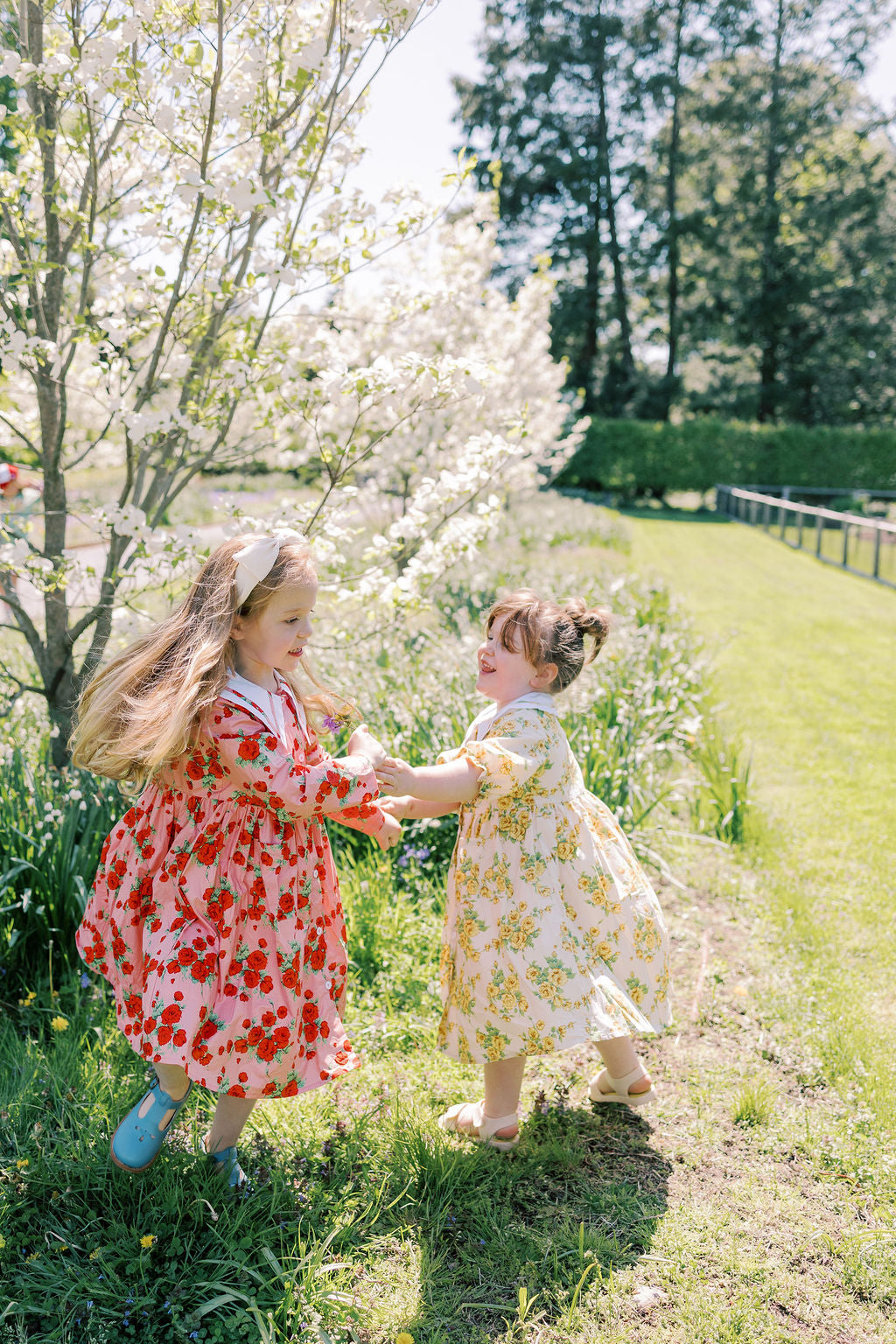 Two young girls in floral dresses standing in a garden with cherry blossom trees.