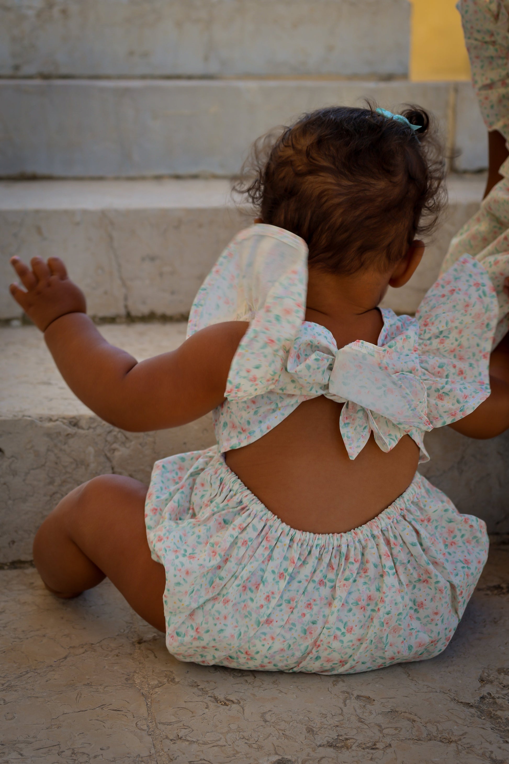 Baby in a floral outfit with a large bow sitting on stone steps.