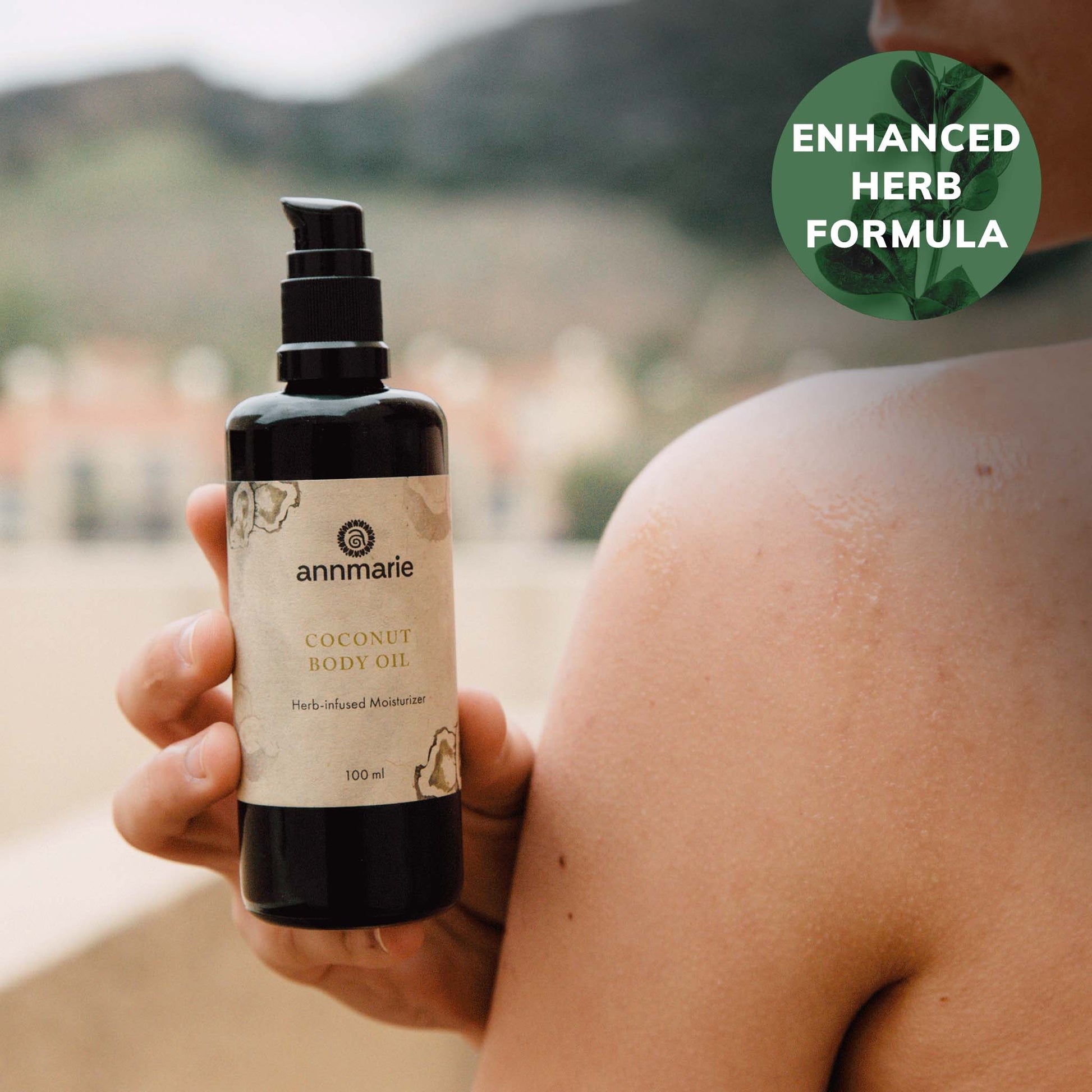 Coconut Body Oil herb-infused moisturizer held next to moisturized shoulder outdoors, 100 ml bottle with enhanced herb formula badge.