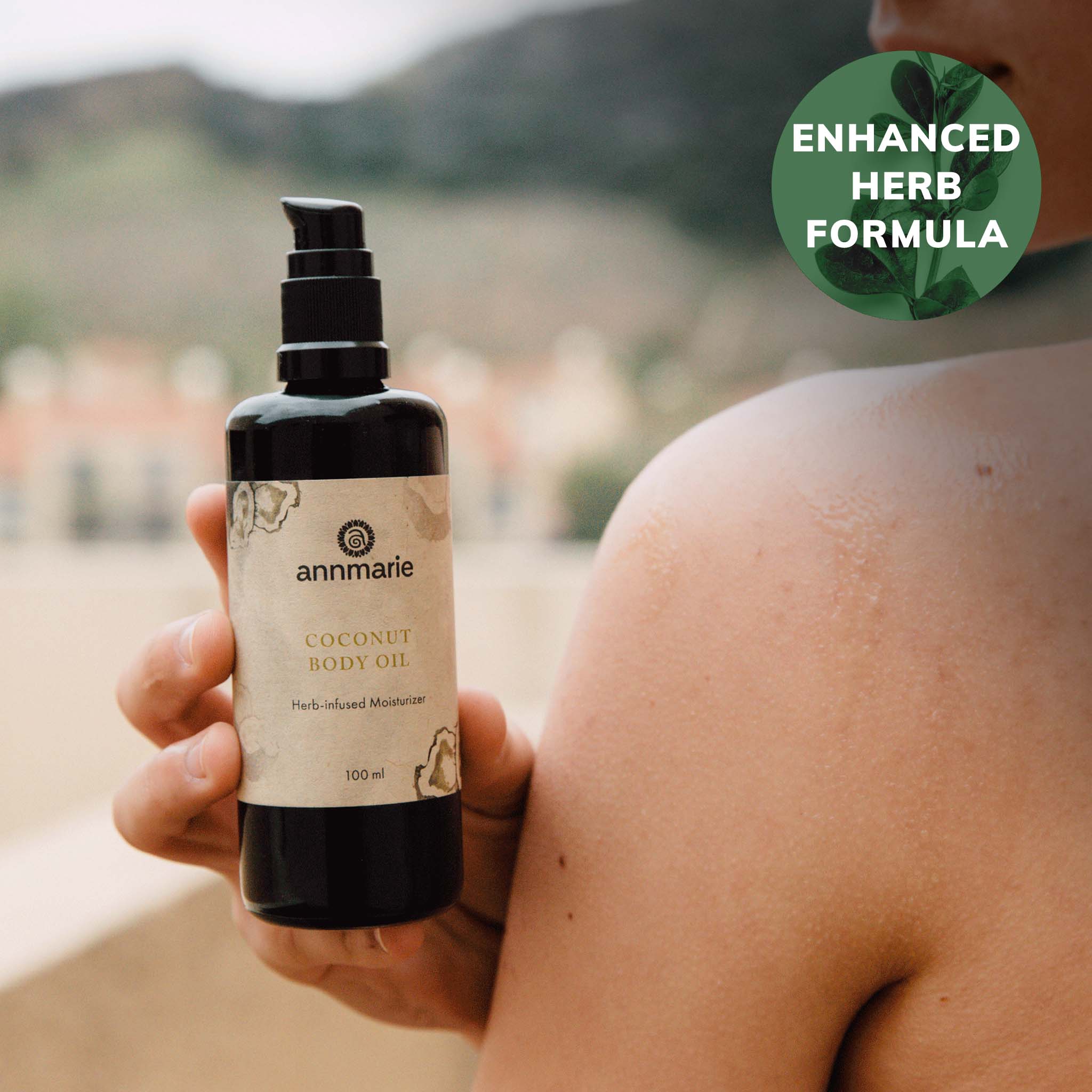 Coconut Body Oil herb-infused moisturizer held next to moisturized shoulder outdoors, 100 ml bottle with enhanced herb formula badge.