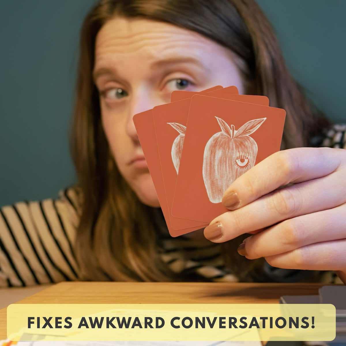 Person holding playing cards with an apple design, text 'FIXES AWKWARD CONVERSATIONS!' at the bottom.