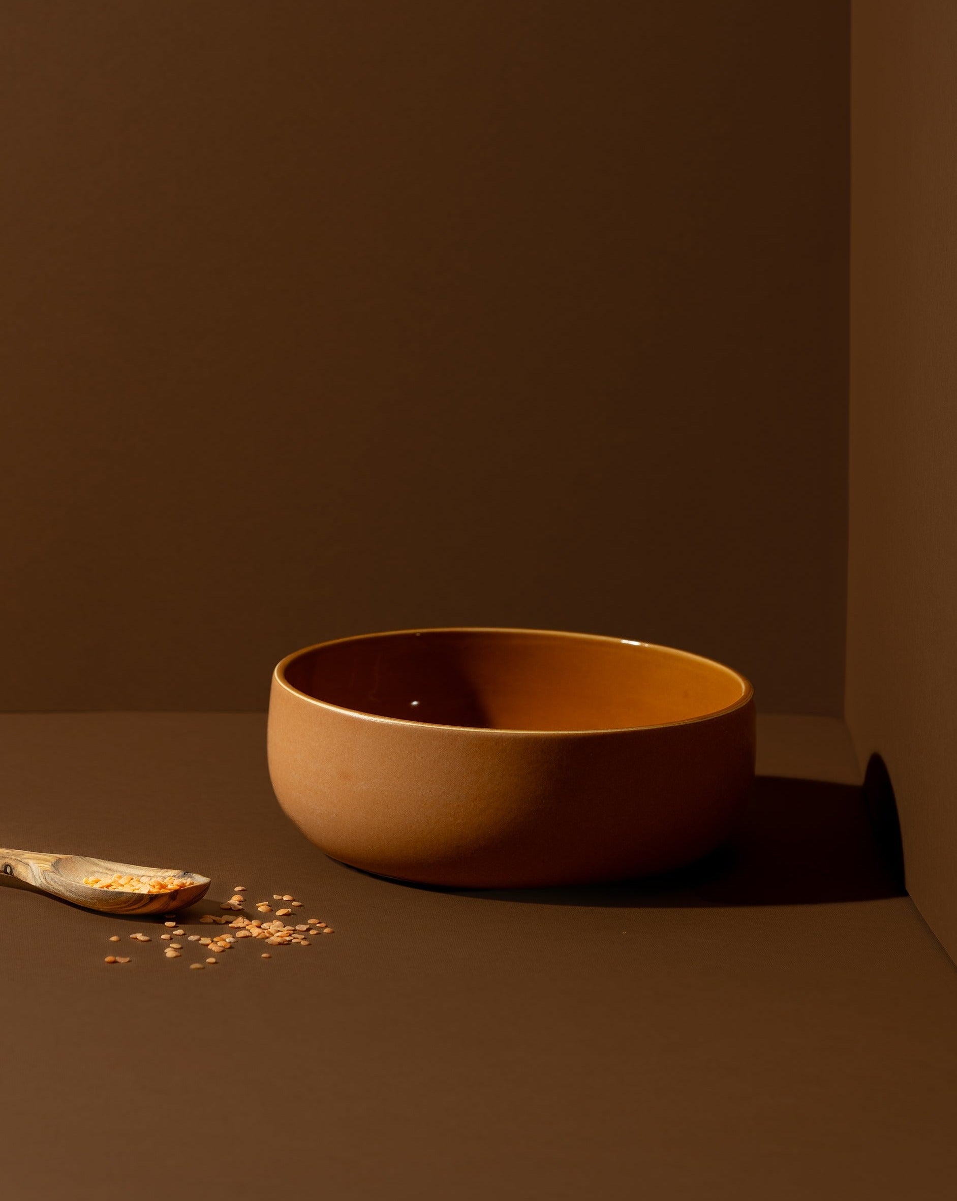 A terracotta-colored stoneware serving bowl placed on a brown surface. A wooden spoon rests beside it, with a few grains scattered around for a rustic touch.

