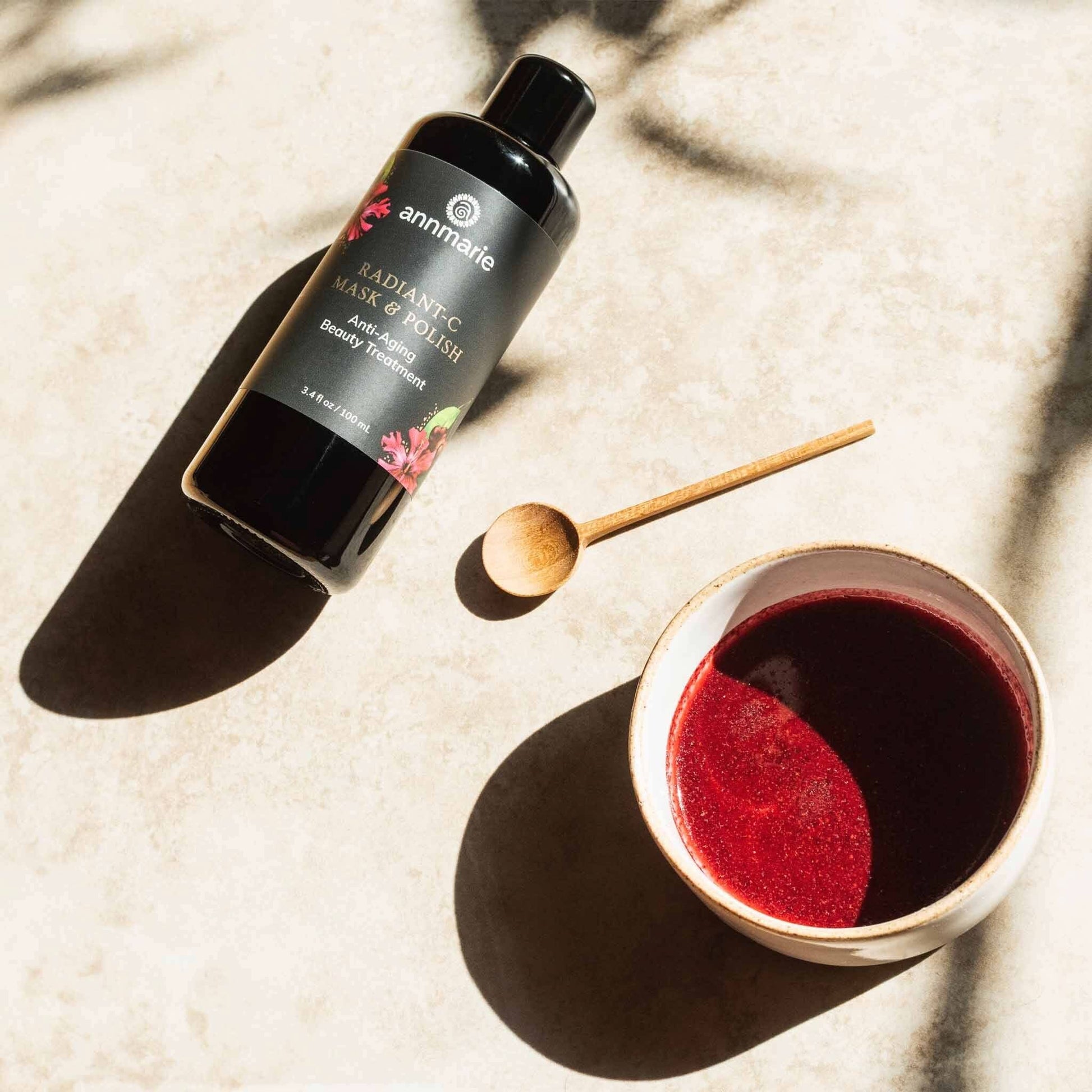Radiant-C Mask & Polish anti-aging beauty treatment bottle styled on a sunlit surface with a wooden spoon and bowl of magenta botanical mixture