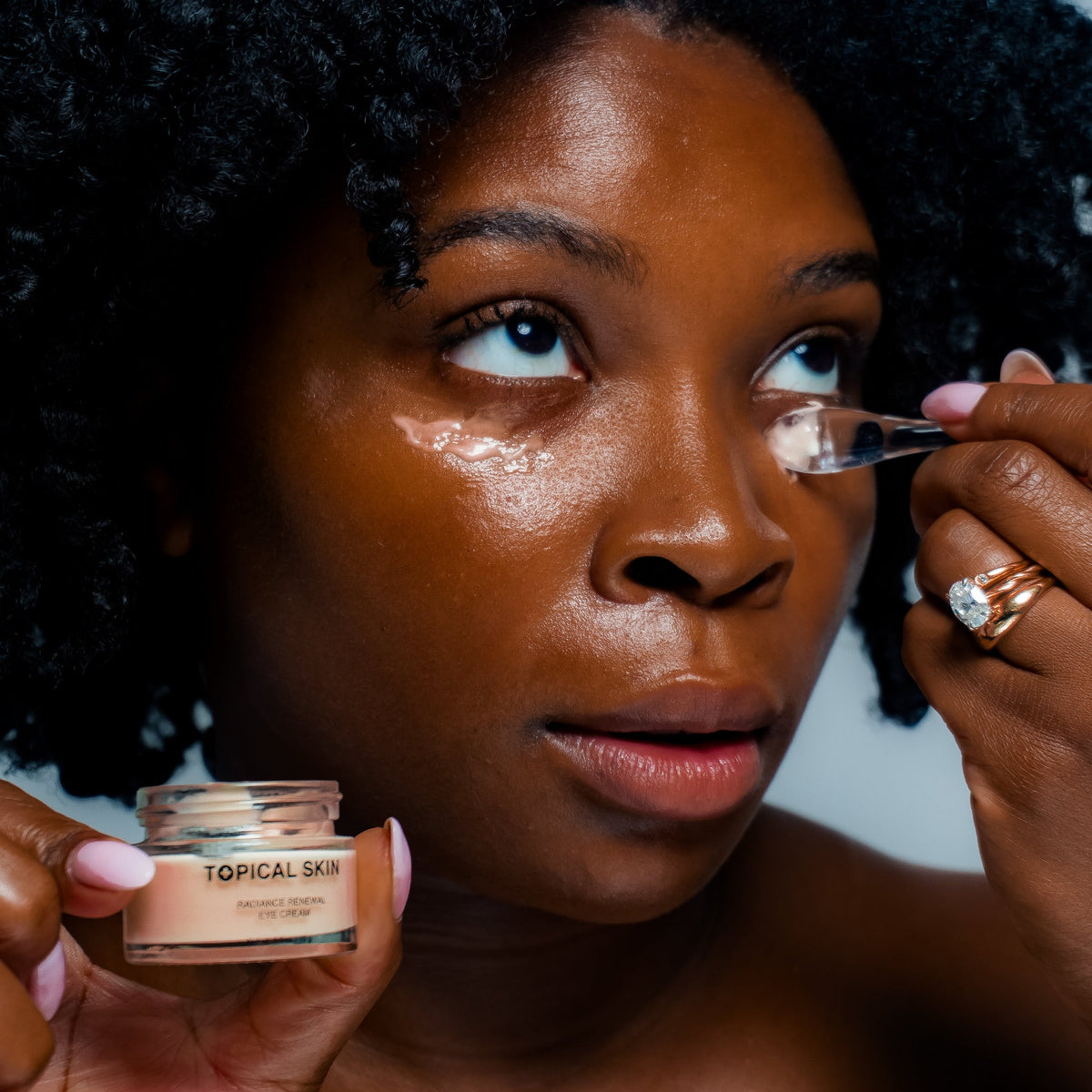 A close-up lifestyle shot of a woman applying under-eye cream from a small jar labeled TOPICAL SKIN. She is holding the open jar in one hand and using a small spatula to apply the cream to her other eye, with her other hand holding a ring nearby.