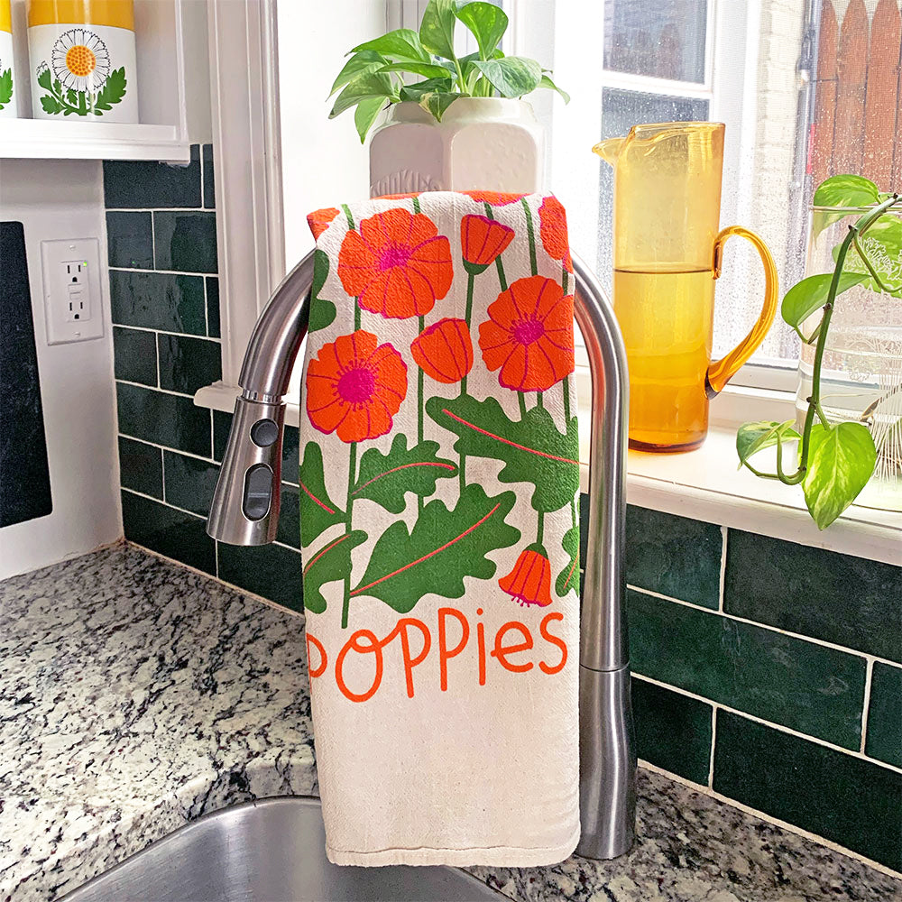 The Neighborgoods Poppies dish towel with a poppy flower illustration hanging over a kitchen sink faucet.