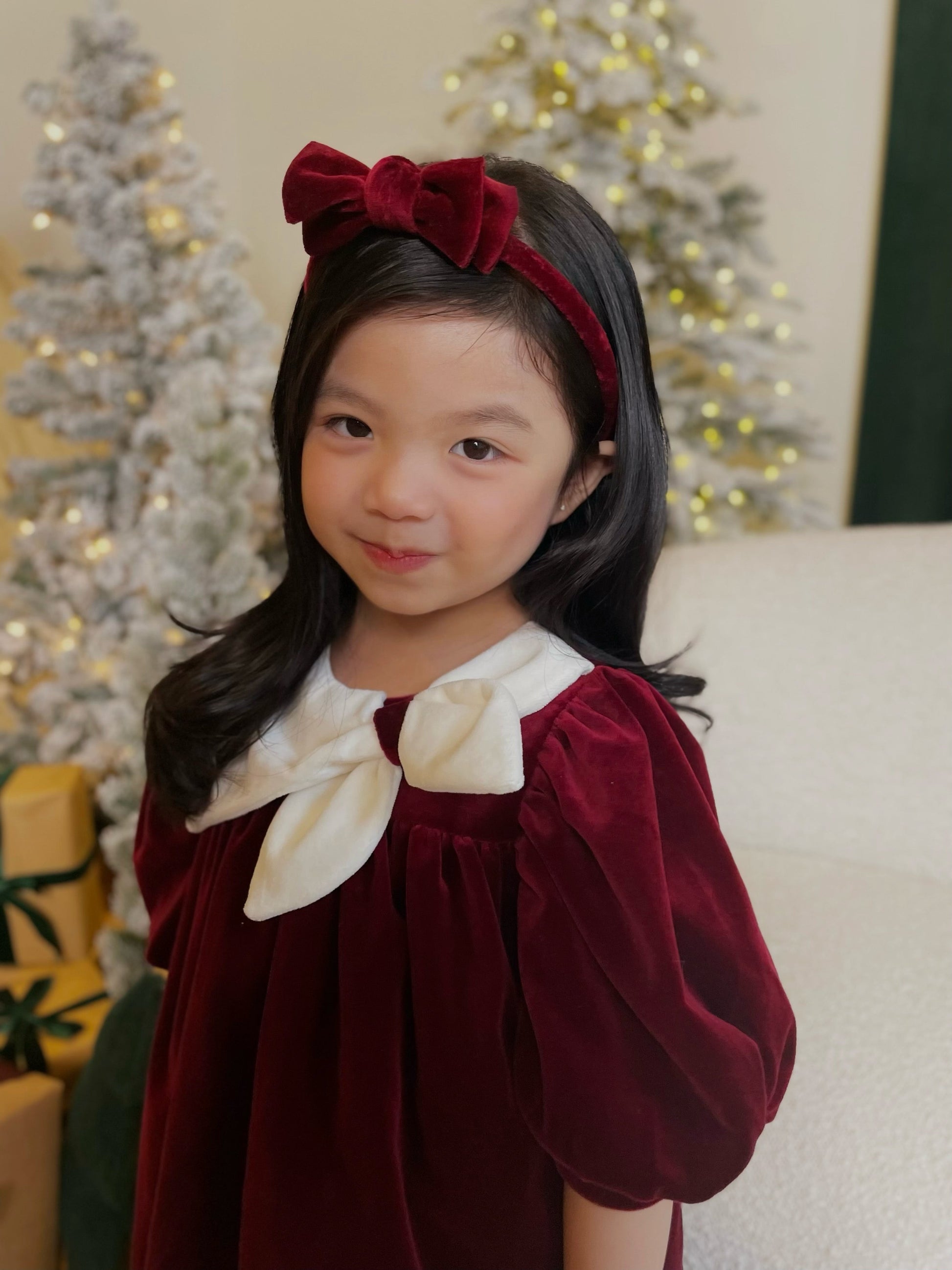 Young girl in a burgundy dress with a white bow and red headband, standing in front of a decorated Christmas tree.