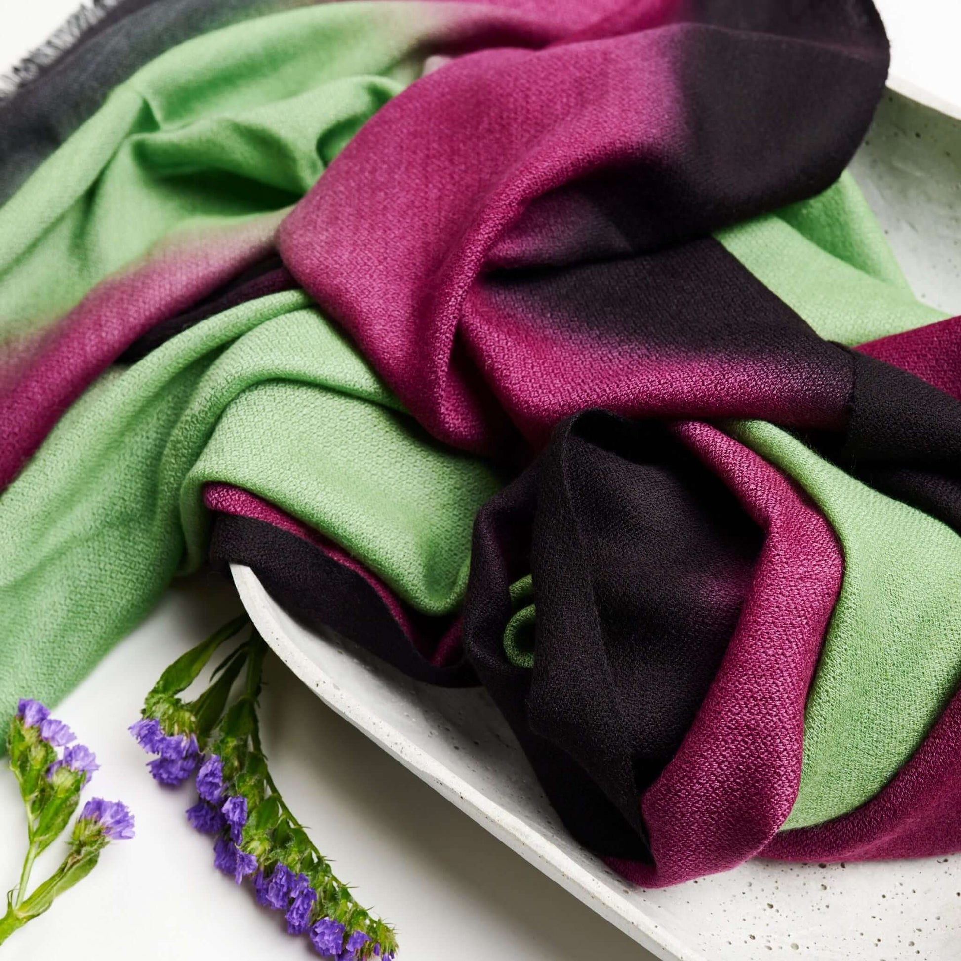 Iris Courtyard Green Purple Hand Woven Cashmere Merino Scarf displayed on a tray with purple flowers.