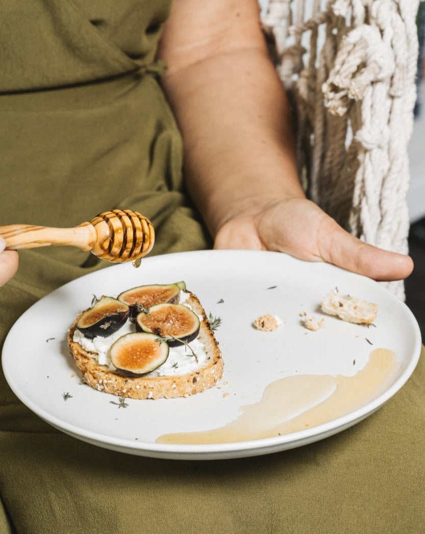 Natural olive wood honey spoon being held by a person, with only the hand and clothing visible, over a plate of food.