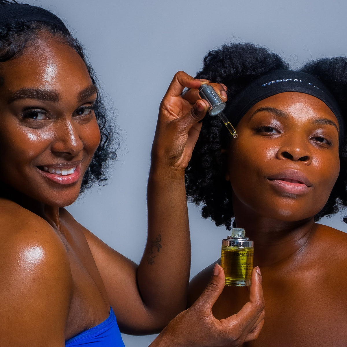Two Black women posing against a solid gray background, one holding a small glass dropper bottle of golden oil while the other receives the drop on her forehead. Both wear black headbands.