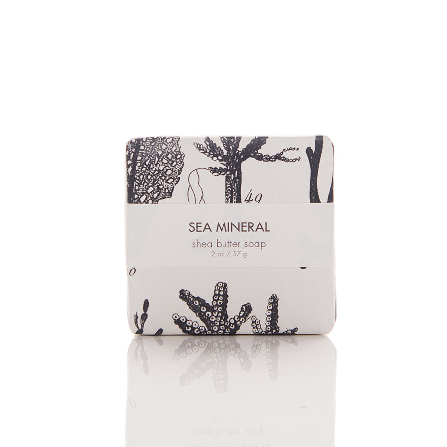 A square sea mineral shea butter soap bar, weighing 2 oz (57 g). The packaging features black and white illustrations of seaweed and coral. The product is labeled with the text "SEA MINERAL shea butter soap.”.