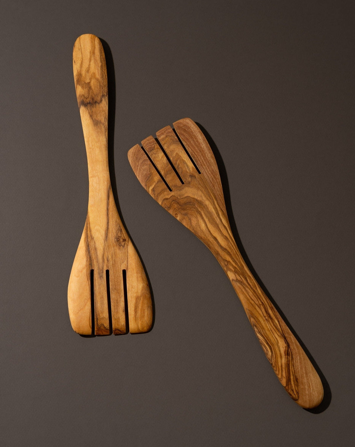 A pair of olive wood serving forks displayed on a dark background, highlighting their smooth finish and natural wood grain.
