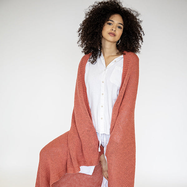 Woman wearing a long rust-pink coloured throw blanket over a white shirt on a plain background
