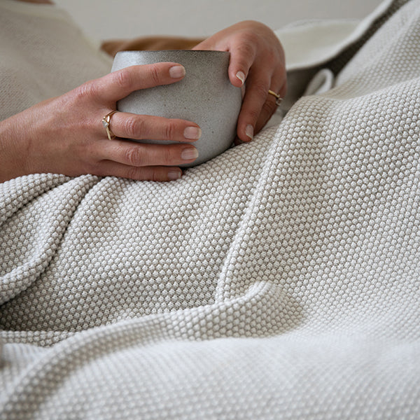 Person holding a ceramic cup with a textured beige throw blanket draped over them.