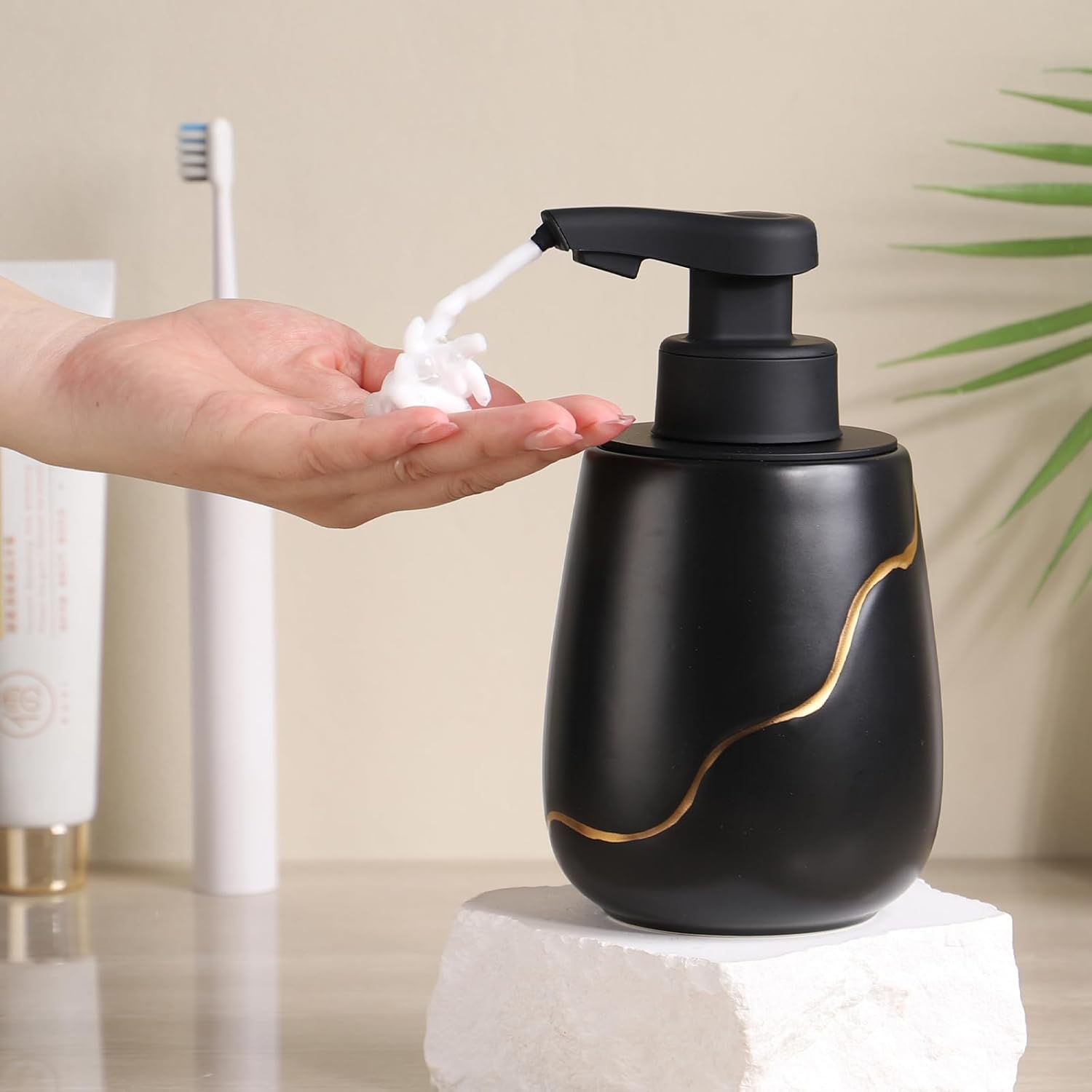 Lifestyle view of a hand receiving foam from the 17 oz BosilunLife matte black and gold touchless soap dispenser. The recycled eco-clay automatic pump is styled on a white stone block on a modern bathroom vanity alongside an electric toothbrush and green plant.