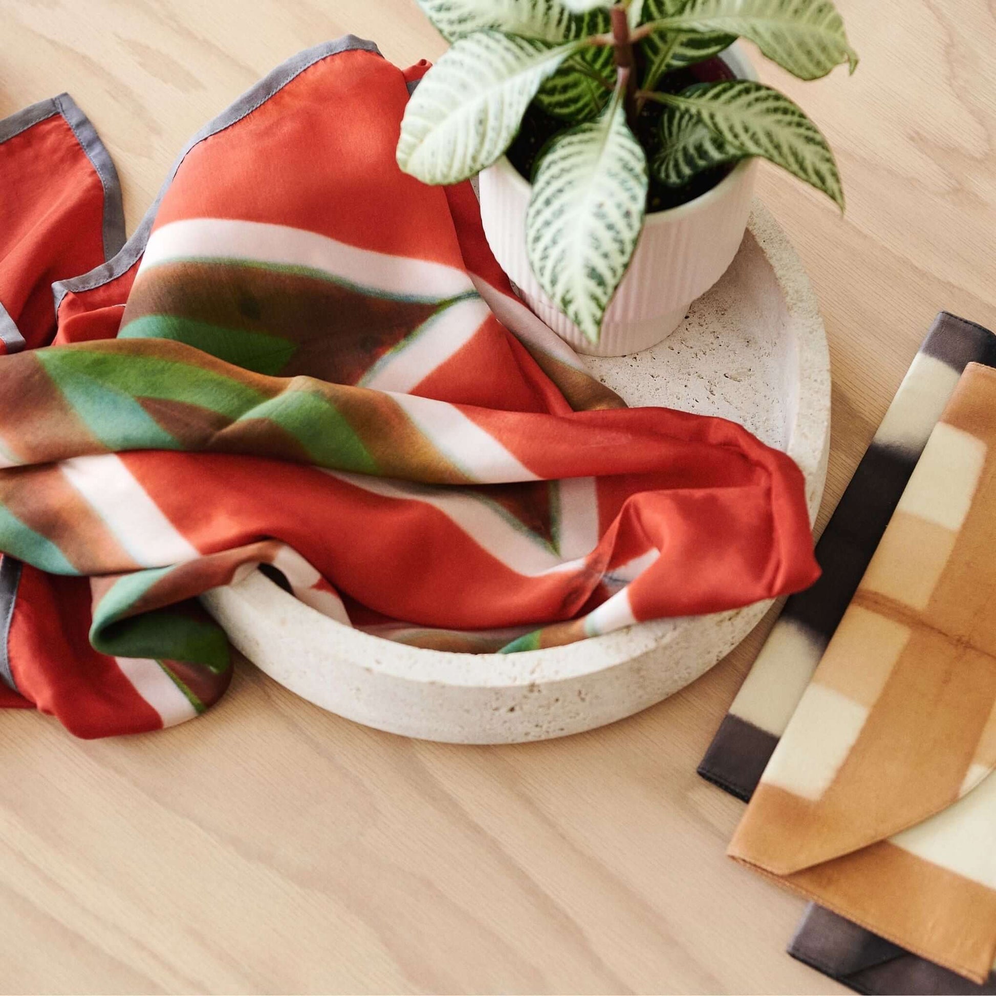 Fawn Square Silk Artisanal Scarf In Multicolor Hand Crafted Shibori Print draped in a bowl with plant decoration.