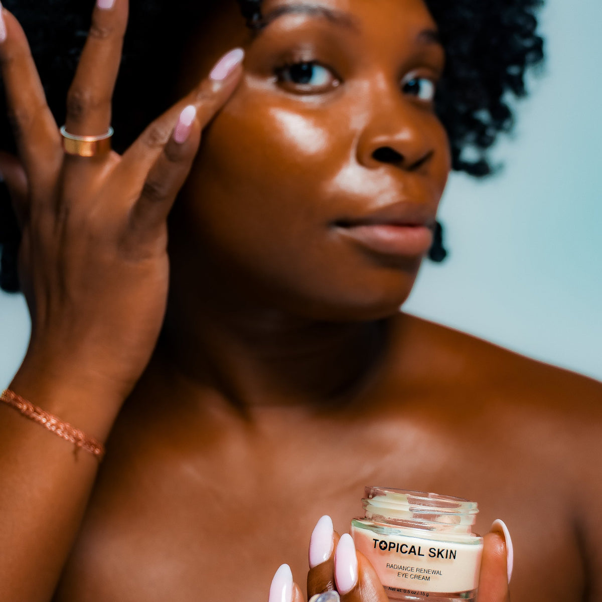 A woman with dark curly hair applies cream to her face while holding a small jar labeled 'TOPICAL SKIN' in her hands. She is wearing a rose gold bracelet and a gold ring, with a light blue background.