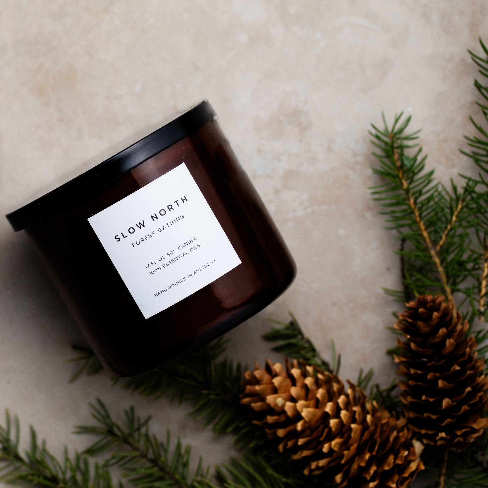 Candle with a label 'Slow North' surrounded by pine branches and cones on a textured surface