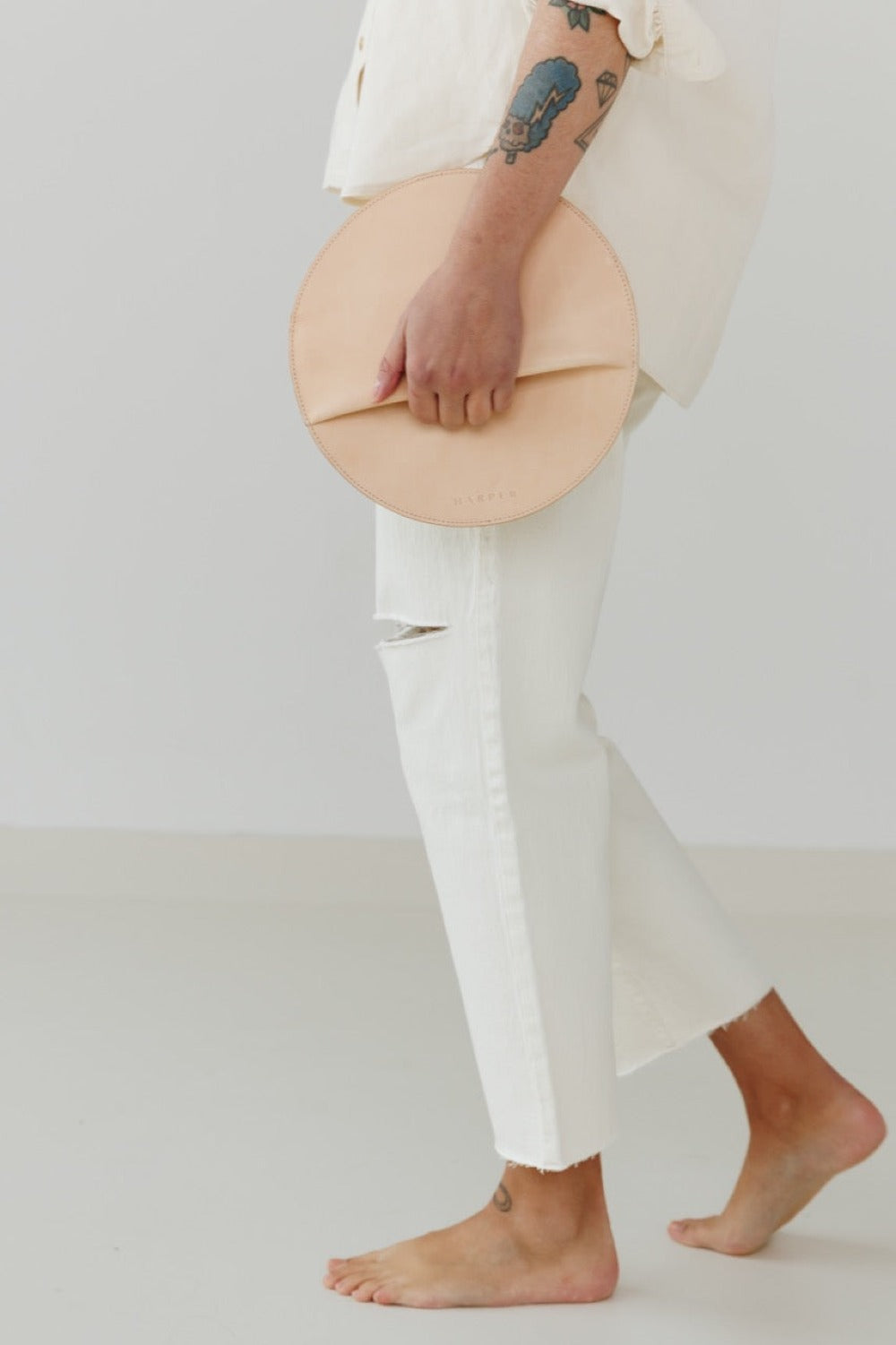 A woman's arms are visible, holding a circular natural leather clutch with the reverse side and zipper visible. The zipper includes a matching zipper pull.