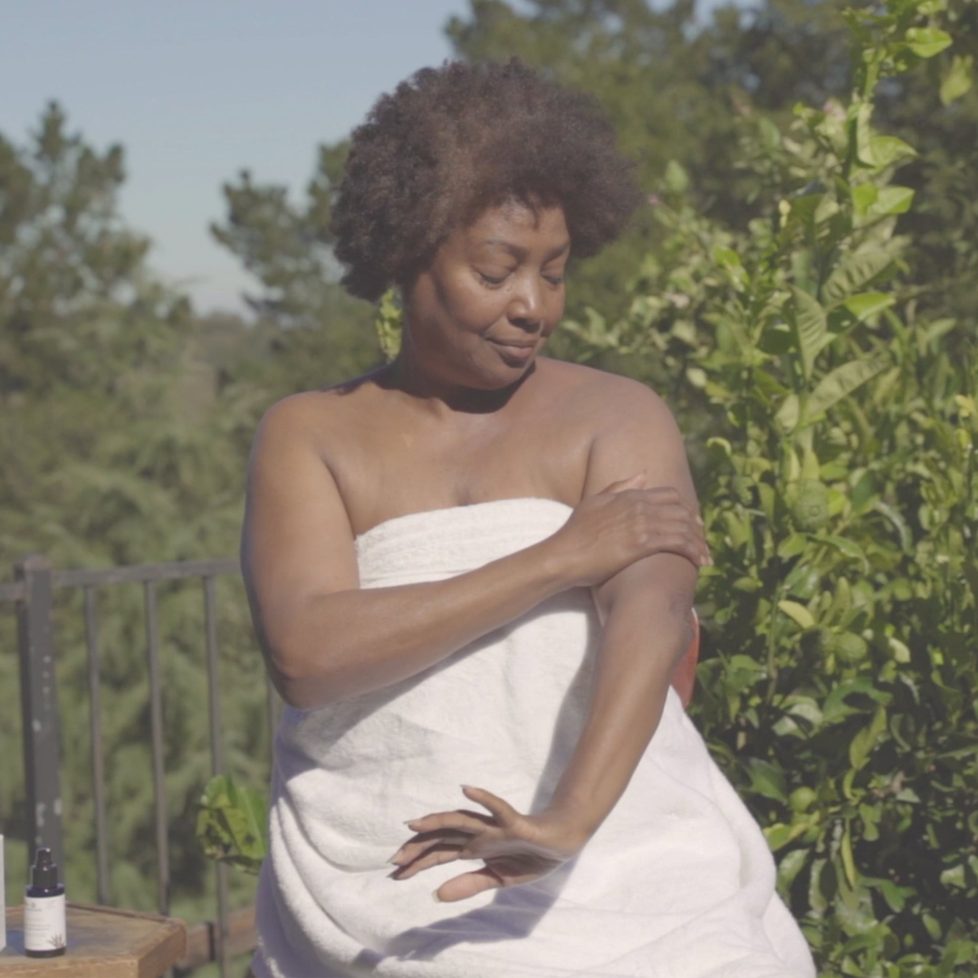 Video of woman outdoors applying Annmarie Sun Love Everyday Sheer Sunscreen SPF 20 to her arm while wrapped in a white towel surrounded by greenery.