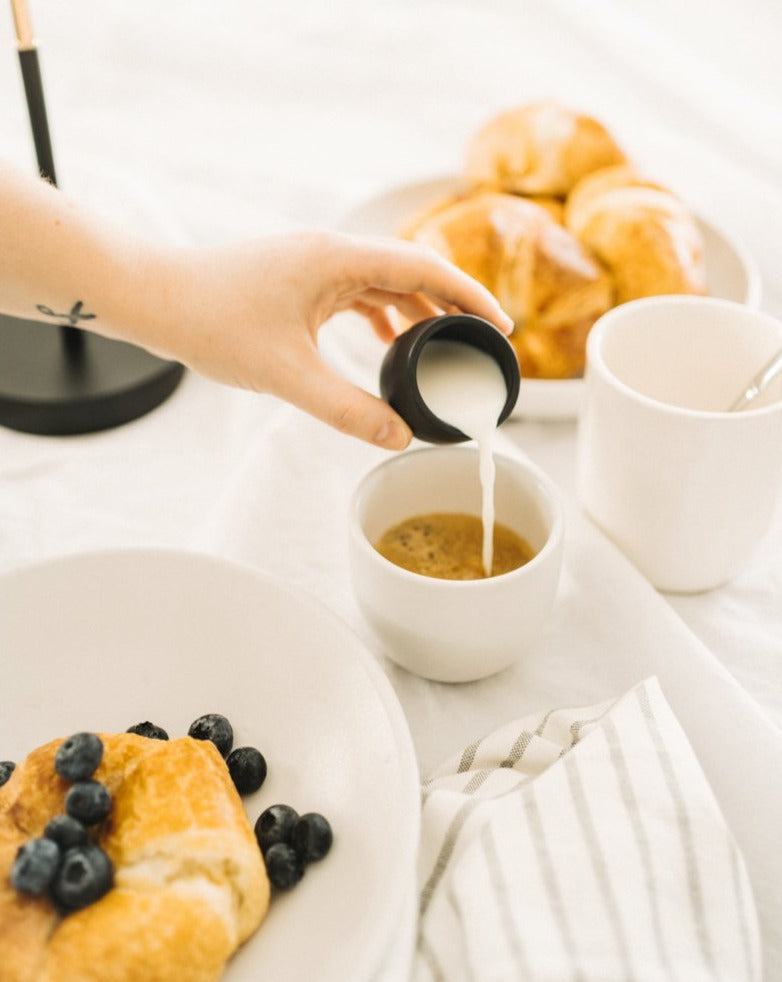 Milk being poured into a stoneware creamer cup, and two plates—one with bread and the other with bread and blueberries."