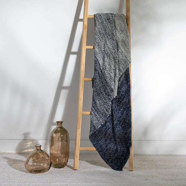 Textured navy and grey cotton throw blanket draped over a wooden ladder with decorative bottles on a light wood floor.