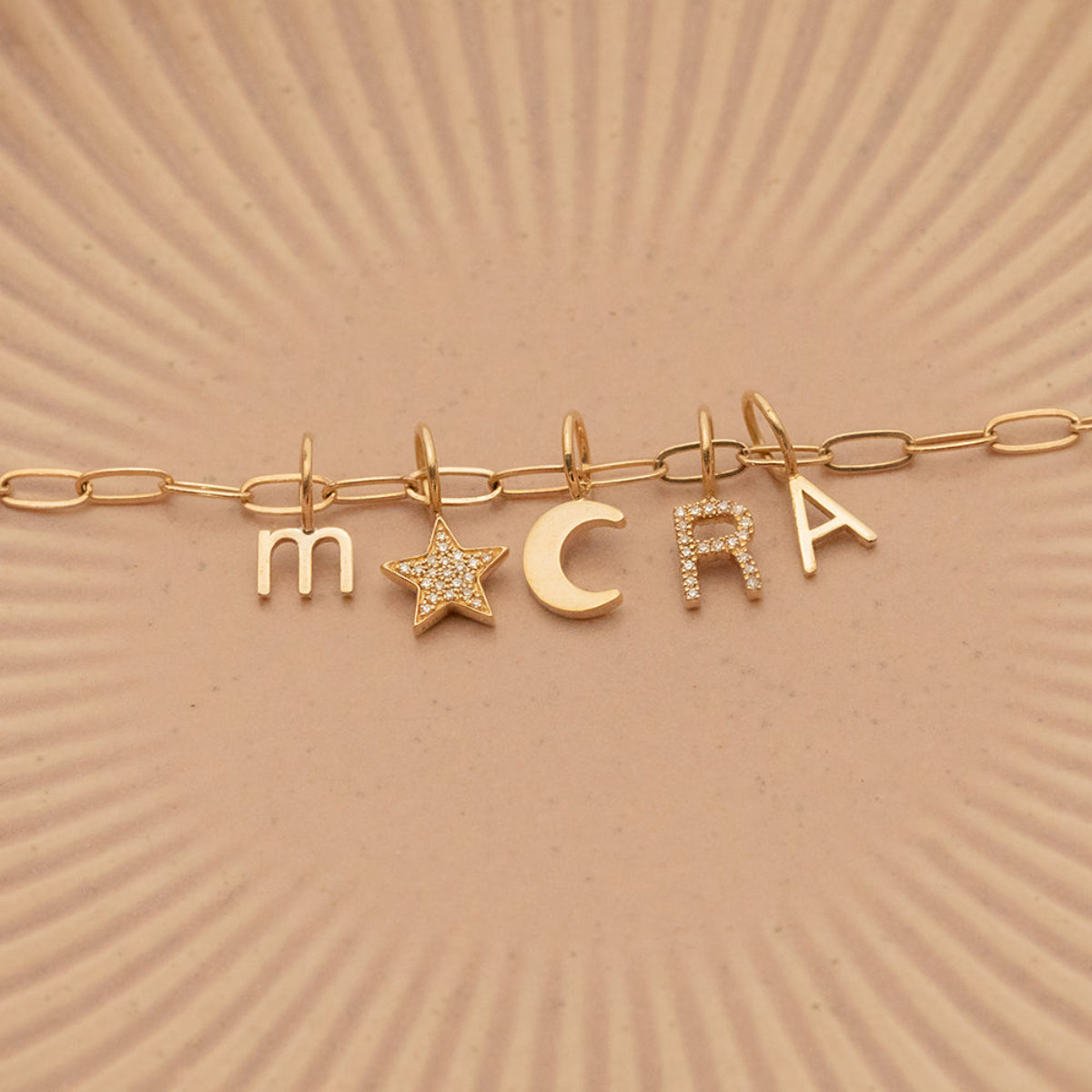 An Element Long Link Chain with a Petite Letter Charm in M, Charm Pendants in Moon and Pavé Star, Pavé Letter Charm in R and Gold Letter Charm in S against a beige background.