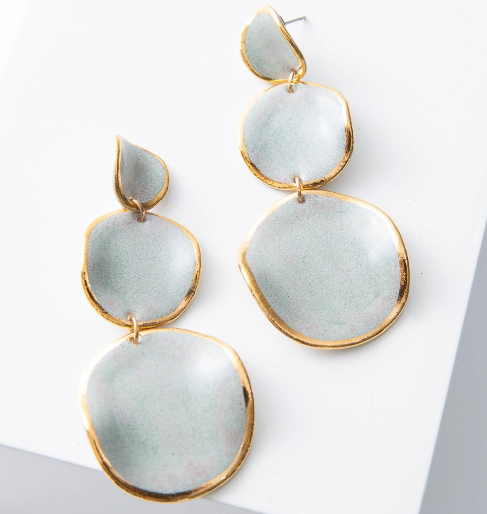 The Stacked Spheres Earrings are dangly earrings composed of three flat, disc-shaped pieces of porcelain stacked on top of each other. The top disc is small, and the discs get larger as you move down the earring. Each disc is composed of light grey porcelain outlined in a ring of shining gold. Rather than being perfectly flat, the discs have a wavy, irregular shape.