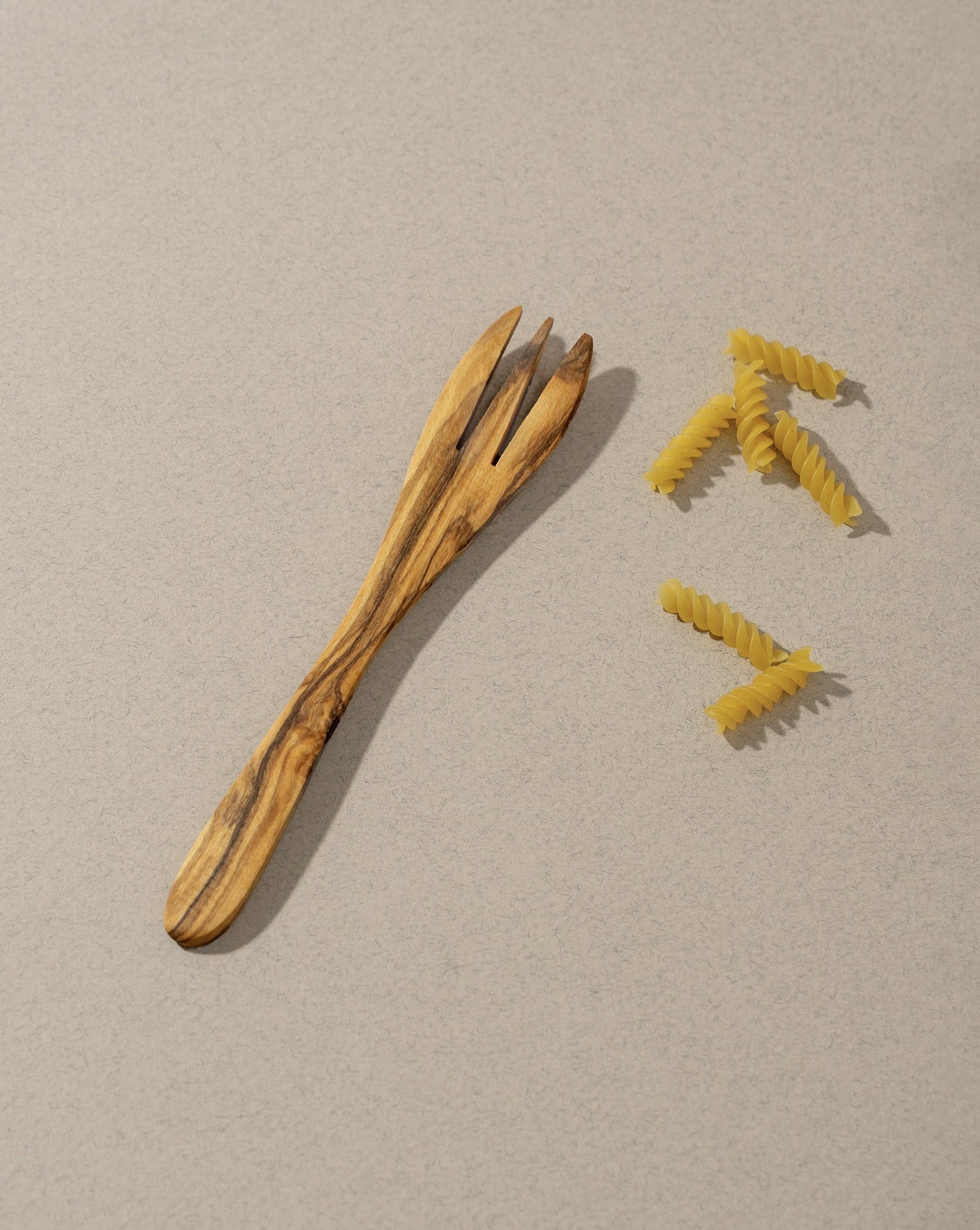 Single natural olive wood tapas fork placed beside a few pieces of pasta on an off-white surface