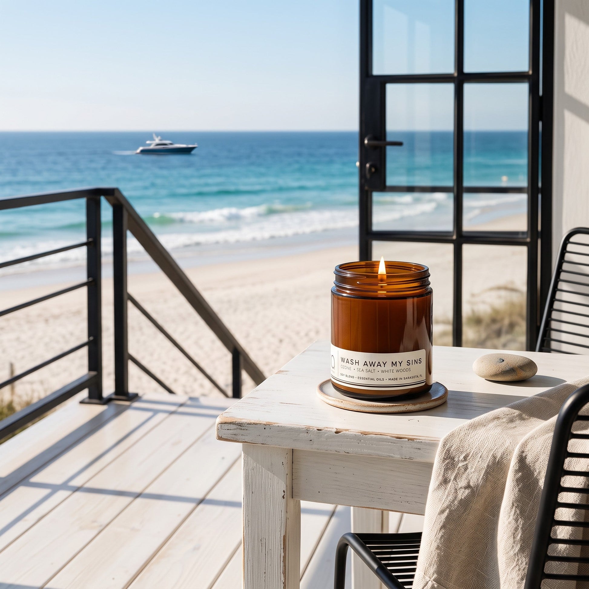 An amber glass jar candle sits on a white wooden table on a beachfront balcony, with a sandy beach, blue ocean, and open black-framed doors in the background.