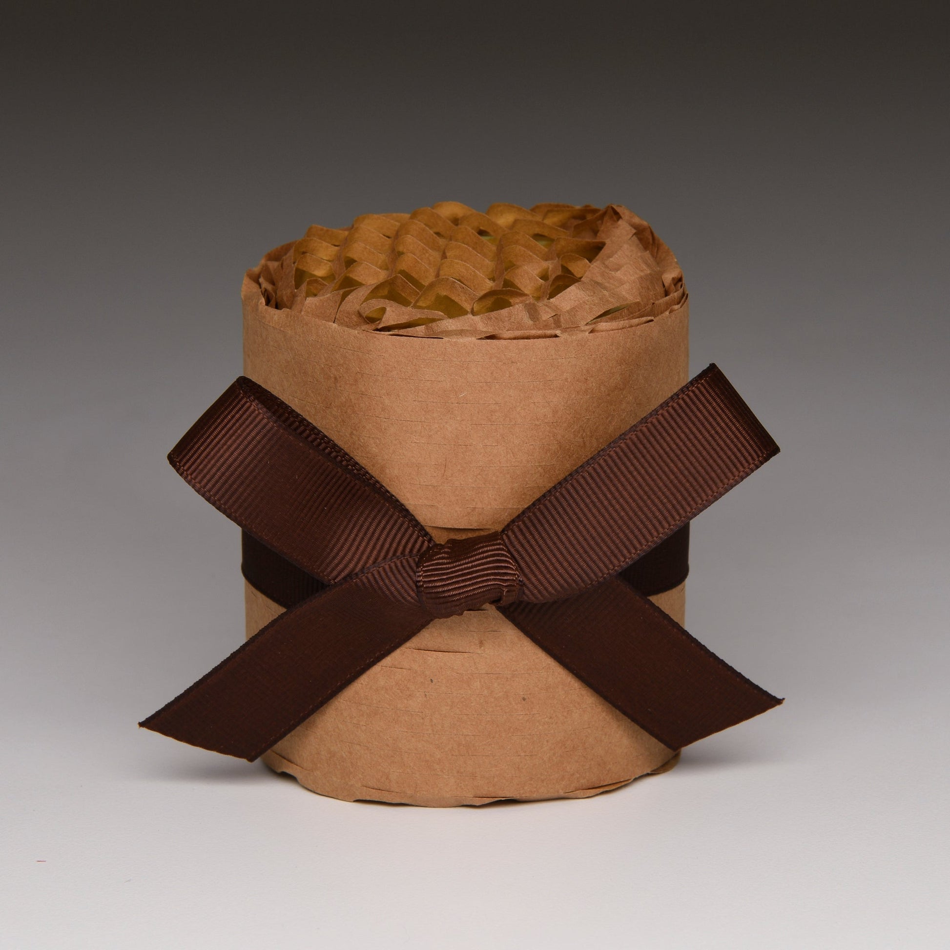 A brown kraft paper cylindrical container filled with crinkled brown tissue paper, tied with a wide dark brown ribbon bow in a studio setting.