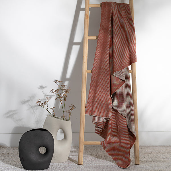 Rust coloured blanket with a ribbed texture draped over a wooden ladder next to a black and white vase.