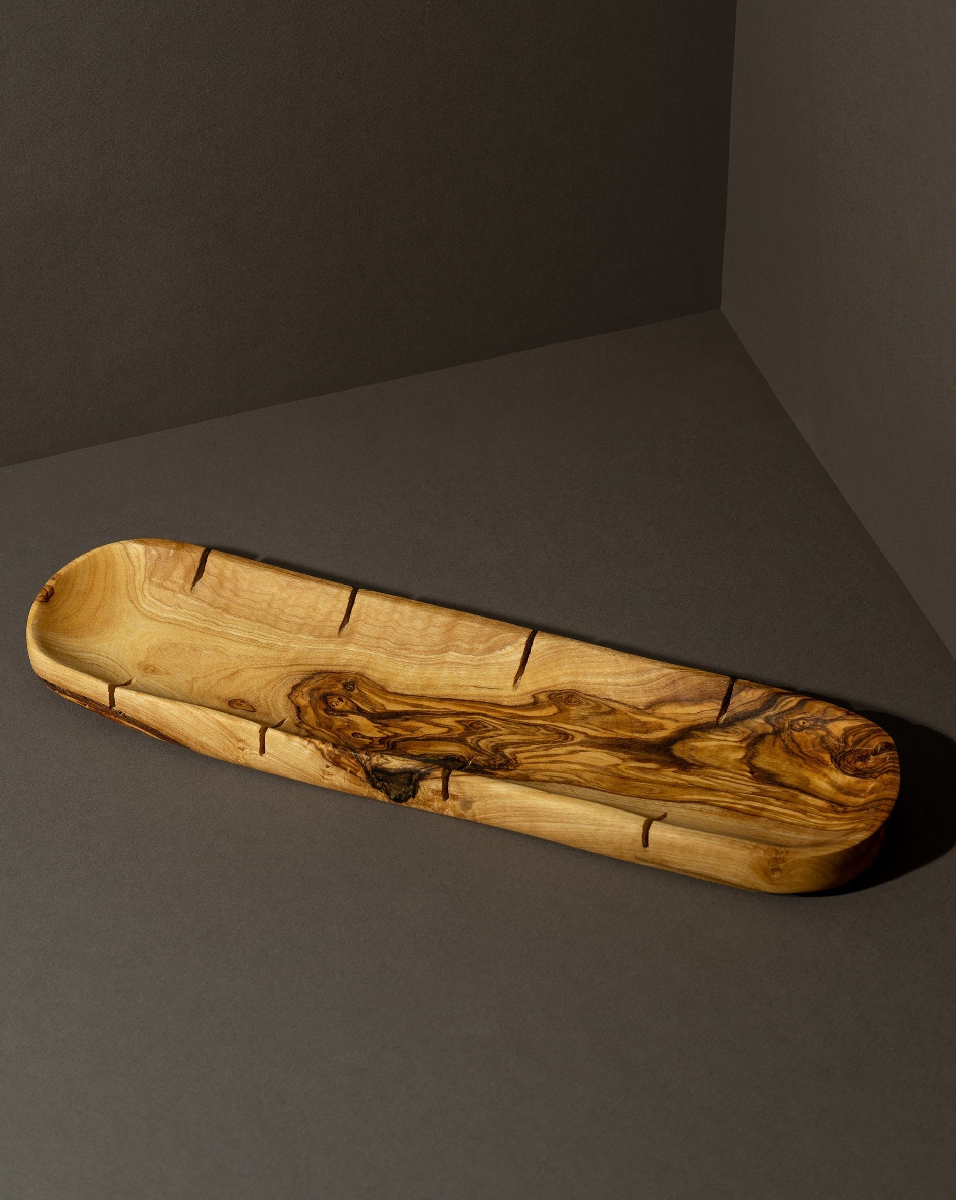 An olive oil bottle and a loaf of bread on a wooden board, with a small wooden bowl of olives beside it, all placed on a white surface.