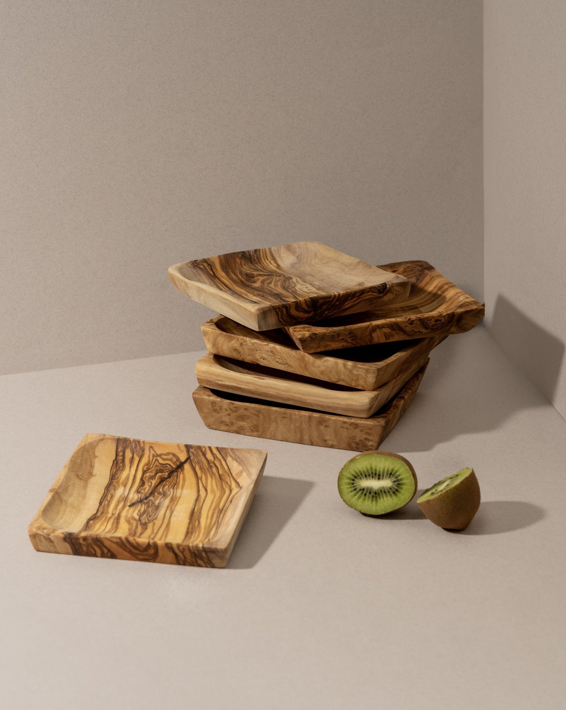 Stacked square olive wood bowls with a kiwi and its slice on a beige surface.
