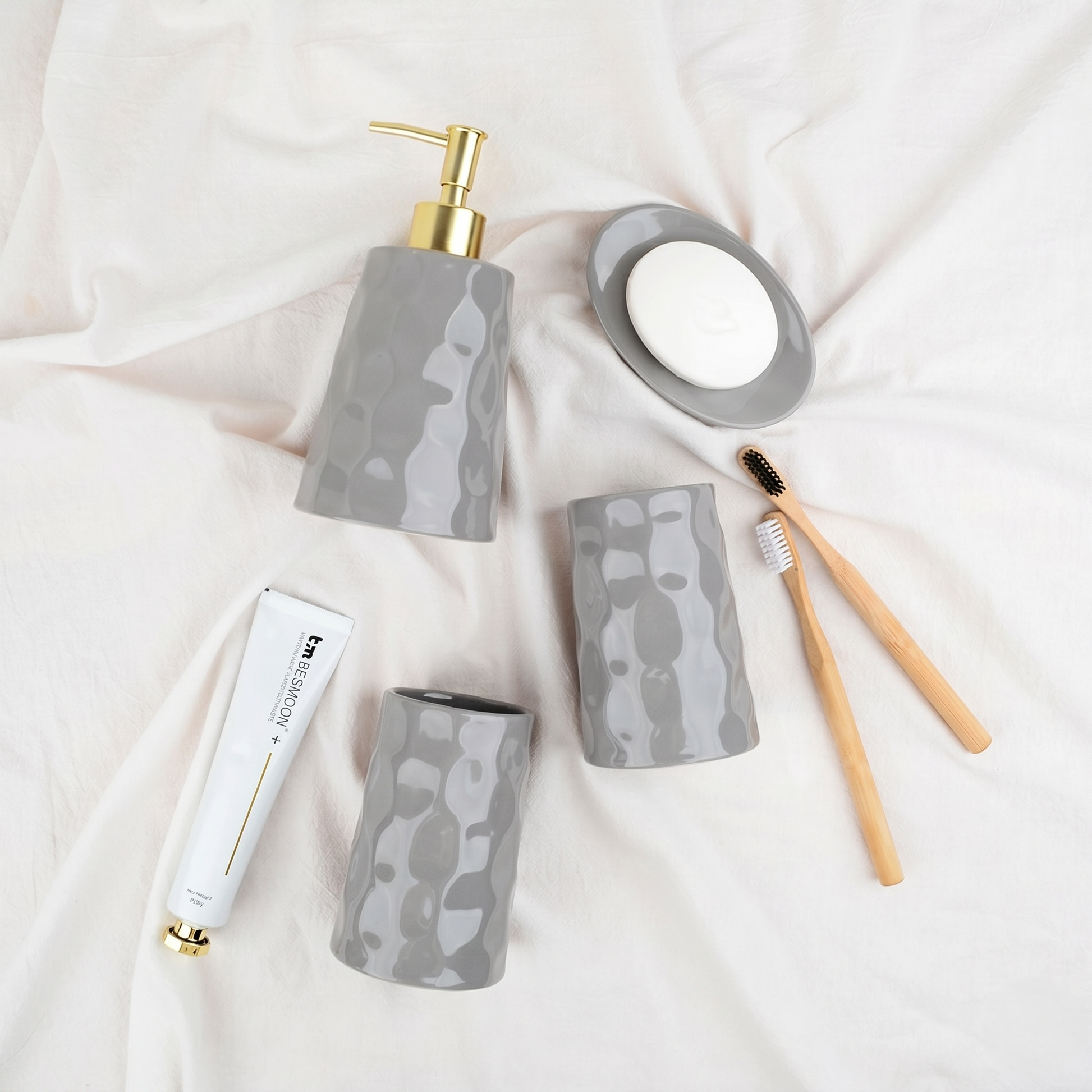 Flat lay lifestyle shot of the BosilunLife Bring Nature Inside 4-piece bathroom accessory set in glossy grey recycled eco-clay. Styled on soft, draped white linen alongside bamboo toothbrushes, the collection features an organic, rippled texture across a liquid soap dispenser with a gold pump, two versatile tumblers, and a matching soap dish, evoking a serene, spa-like aesthetic.
