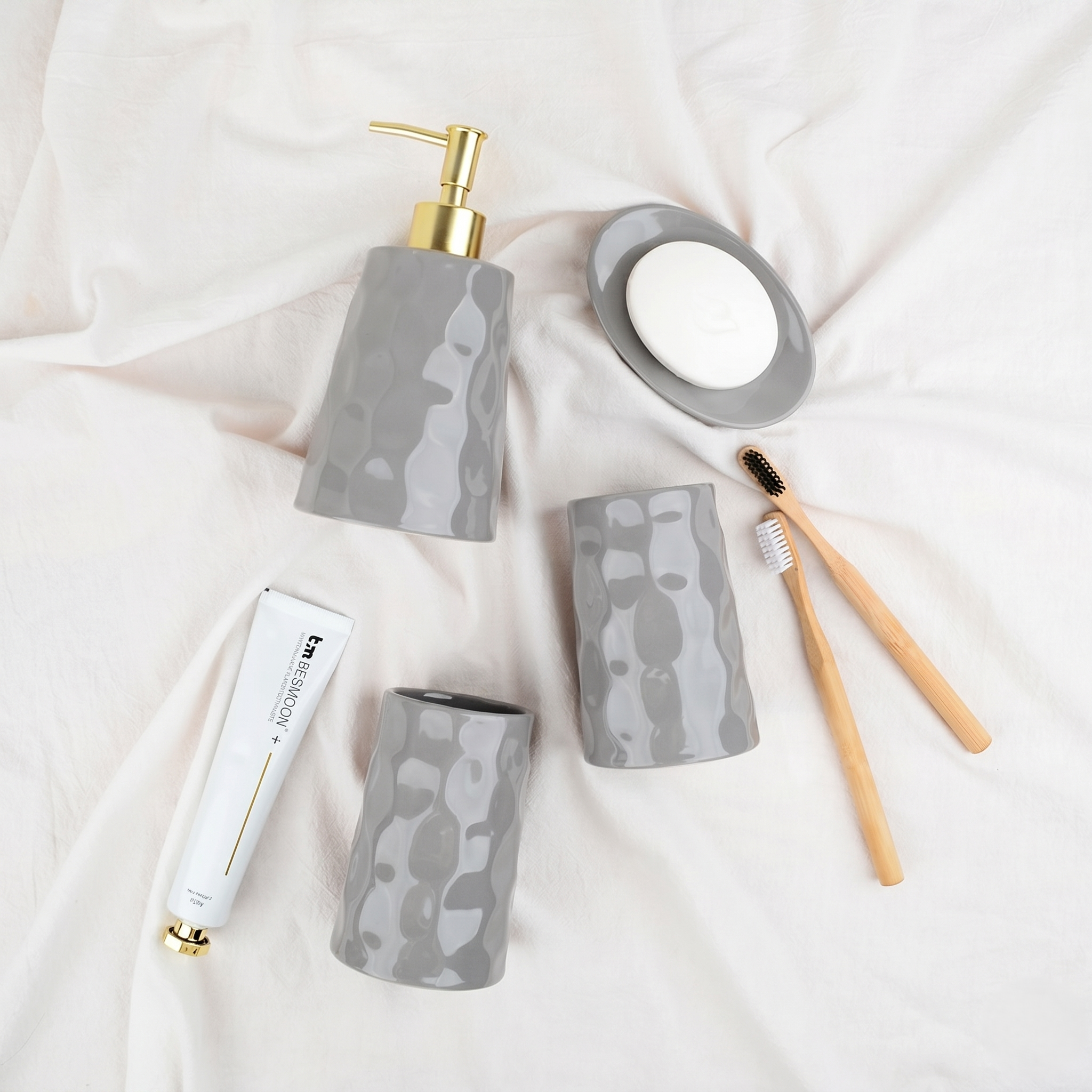 Flat lay lifestyle shot of the BosilunLife Bring Nature Inside 4-piece bathroom accessory set in glossy grey recycled eco-clay. Styled on soft, draped white linen alongside bamboo toothbrushes, the collection features an organic, rippled texture across a liquid soap dispenser with a gold pump, two versatile tumblers, and a matching soap dish, evoking a serene, spa-like aesthetic.
