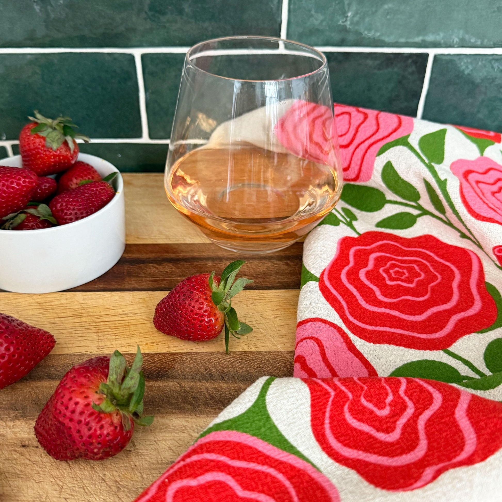 The Neighborgoods Roses dish towel featuring a rose illustration, laid next to freshly cut strawberries and a glass of wine on a kitchen countertop.