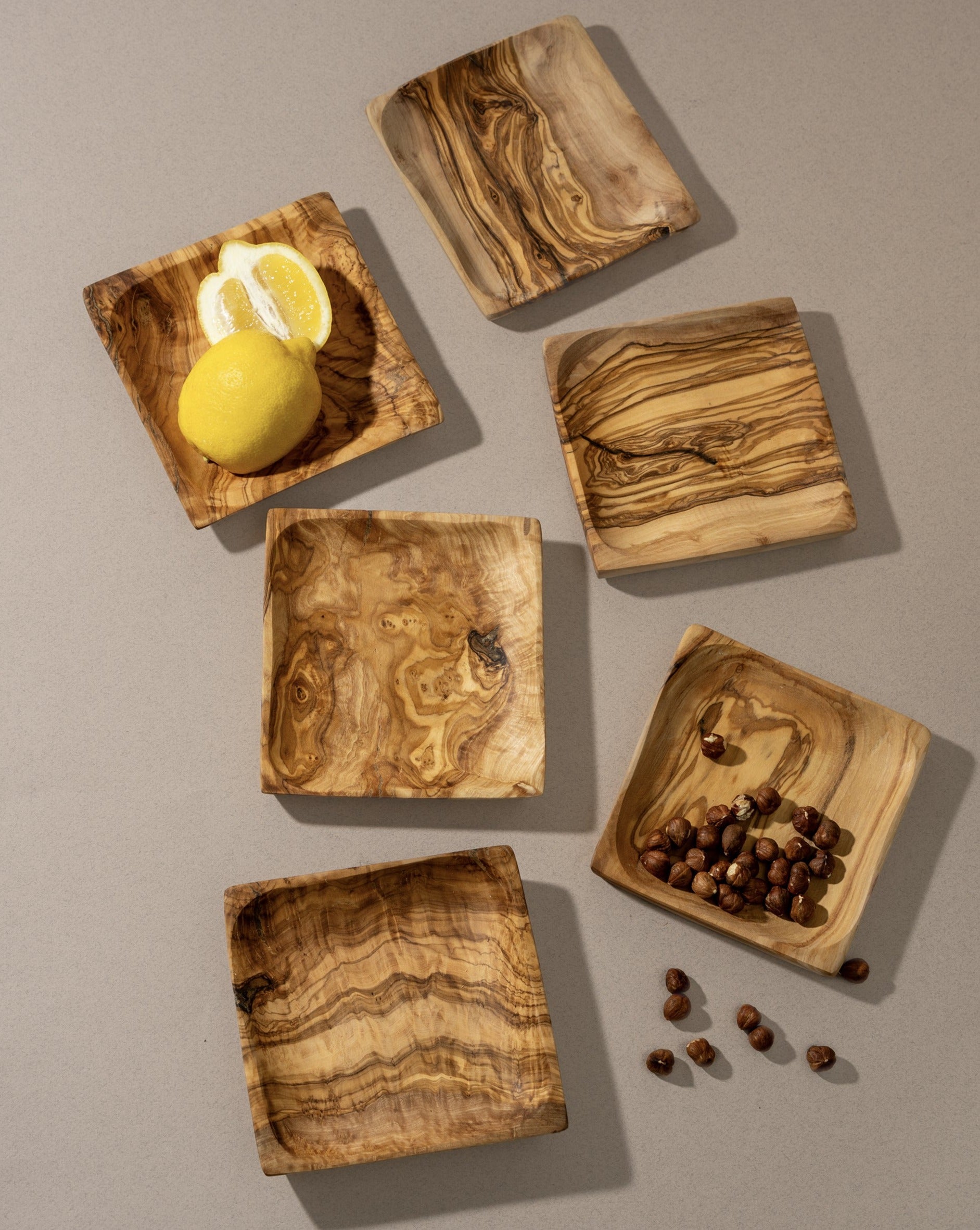 Six square olive wood bowls neatly arranged on a beige surface. Two of the bowls are filled—one with a lemon and the other with coffee beans—while the rest are empty, showcasing their natural wood grain and handcrafted finish.
