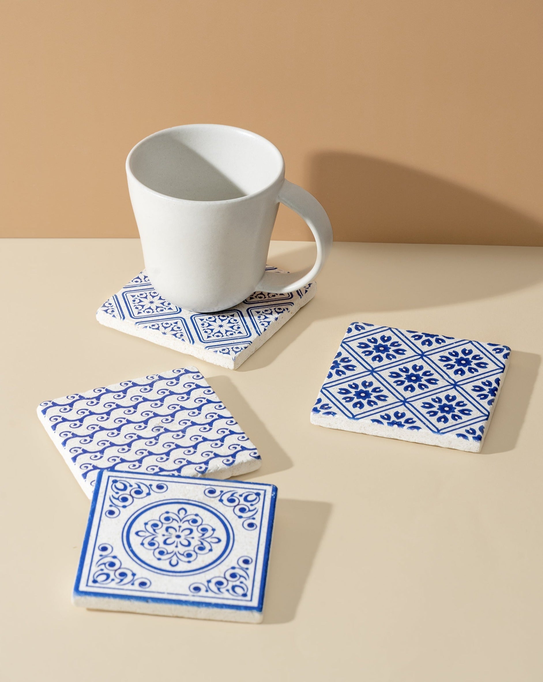 Four square white marble coasters with blue patterned designs, displayed on an off-white background. A cup is placed on one of the coasters, showcasing both style and functionality.