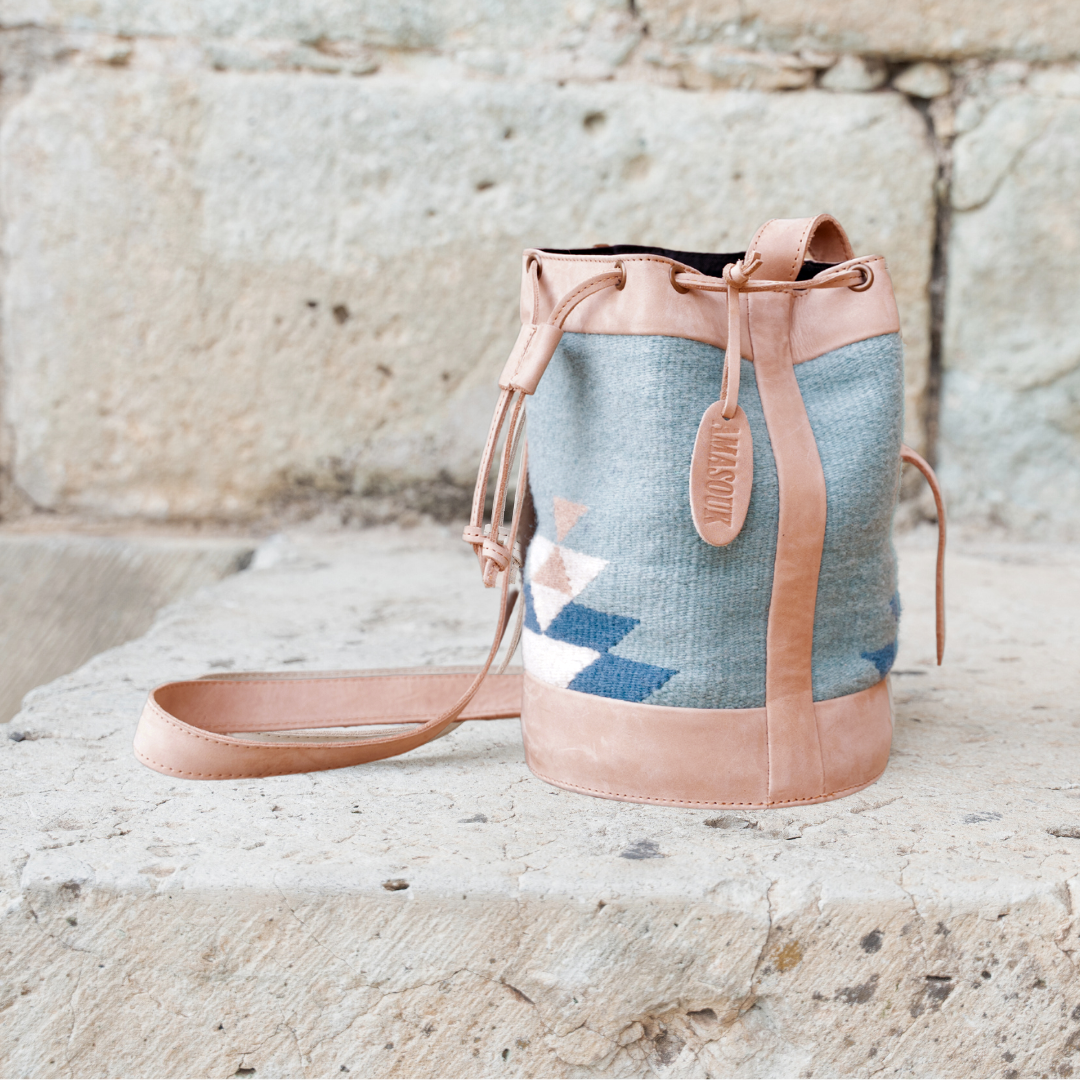 Handwoven blue wool and leather cinched bucket bag with adjustable strap, featuring a soft silhouette inspired by river currents; artisan-made and spacious.