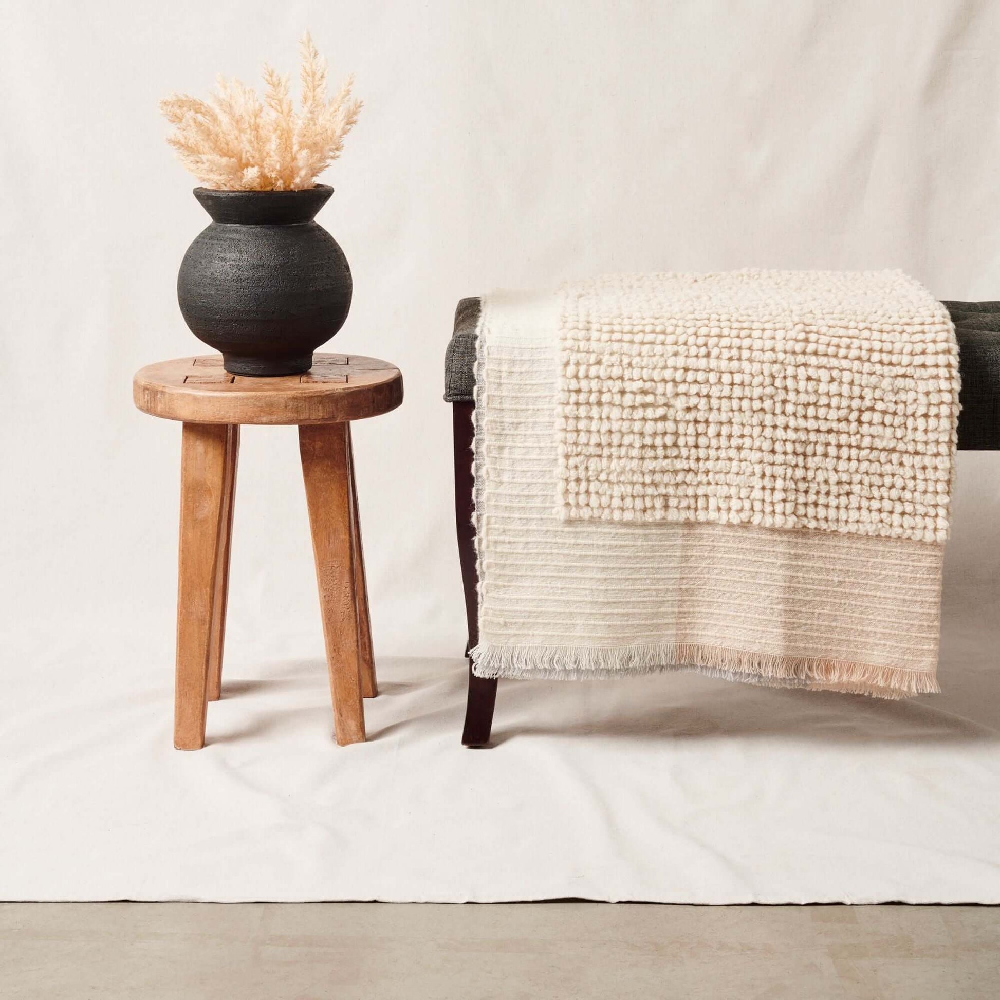 Butterscotch White Handloom Textured Plush Merino Throw displayed on a bench beside a wooden stool and vase with decorative plant.