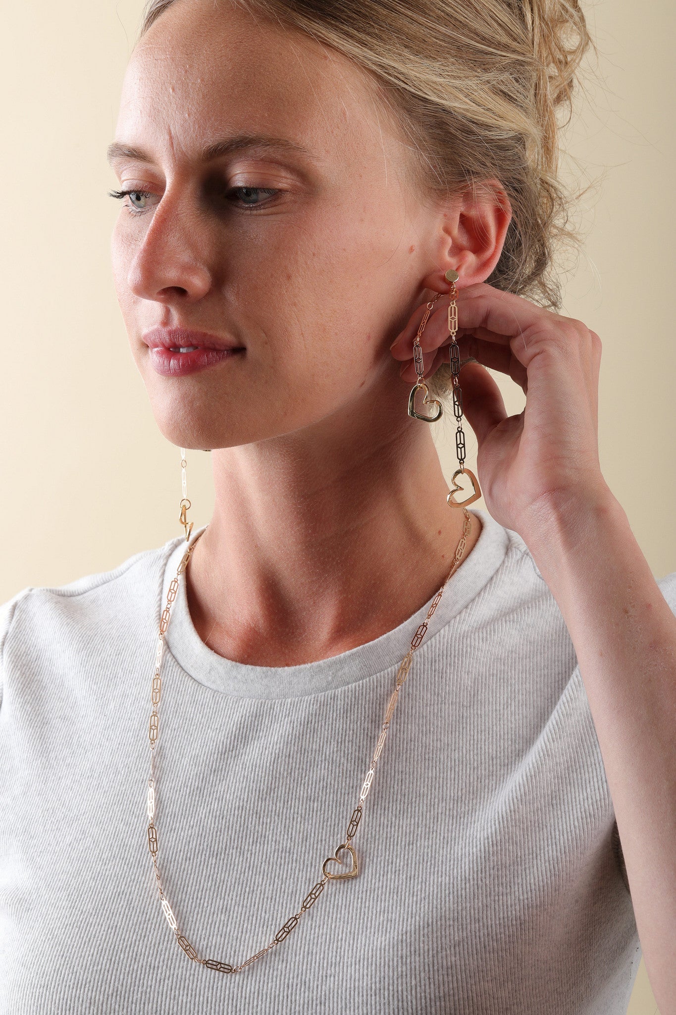 Woman wearing gold heart-shaped earrings and a matching necklace against a beige background