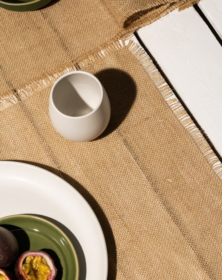 A beige burlap placemat on a gray surface, partially showing a plate with a small amount of food, along with a small cup placed above it. The setup creates a simple, rustic dining arrangement.

