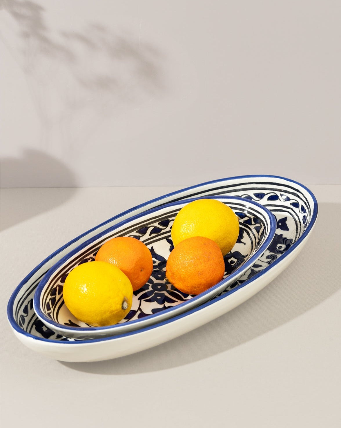 Two oval ceramic plates with blue and white floral designs, stacked on top of each other on a light background. The smaller top plate holds two oranges and two lemons, highlighting the functional and decorative appeal of the set.