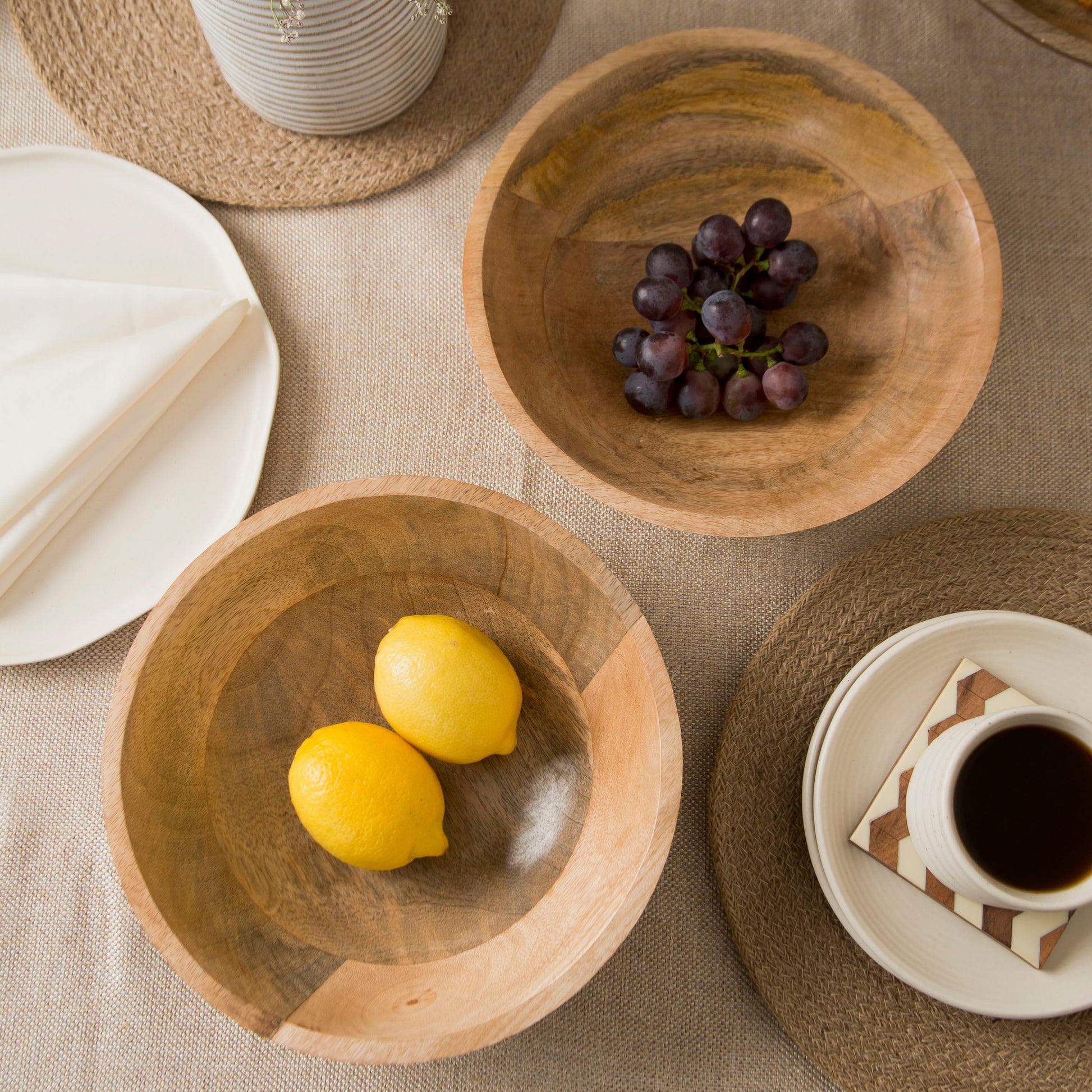 Two natural mango wood bowls containing lemons and grapes on a table with woven placemats.