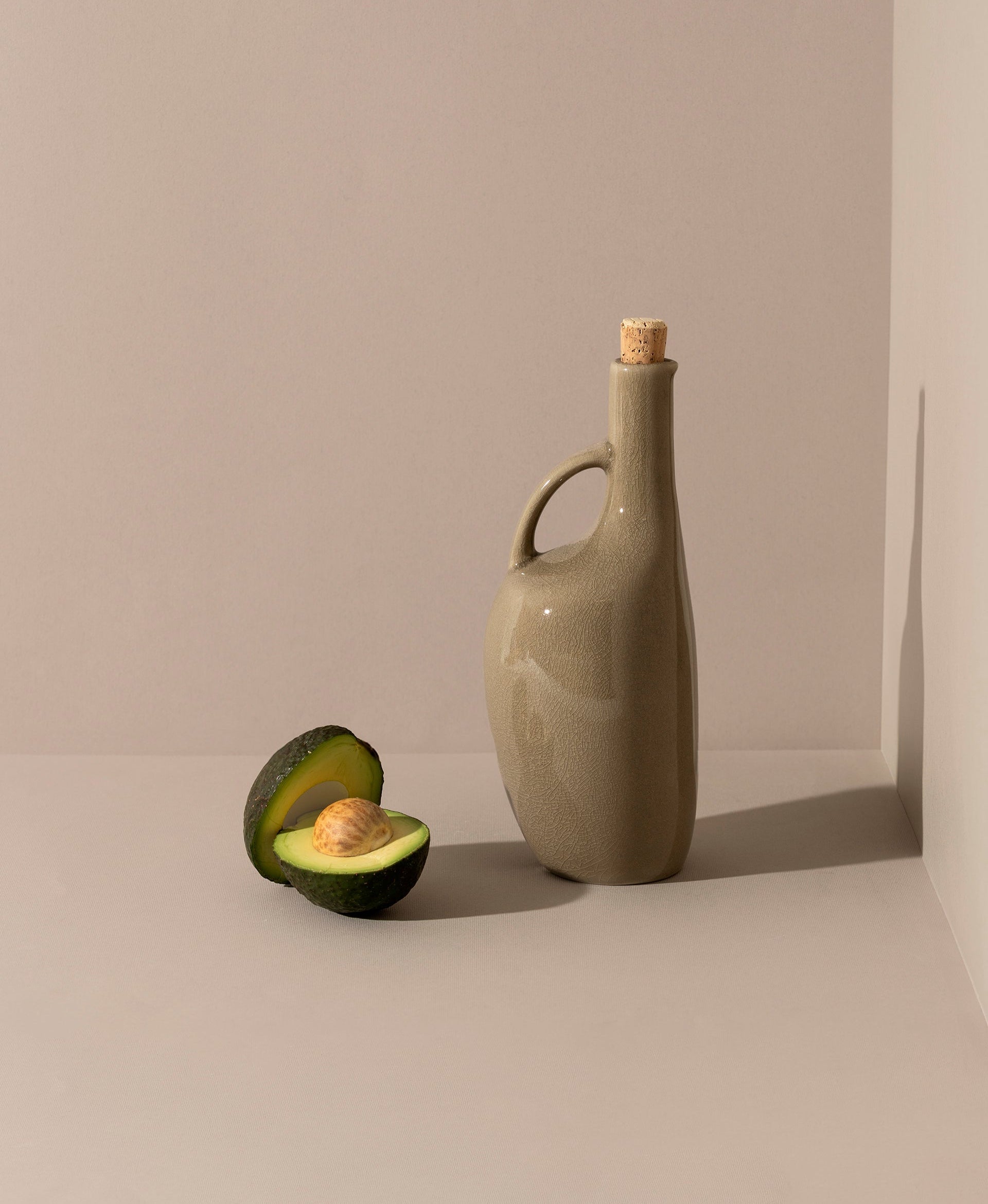 A white ceramic olive oil dispenser with a cork stopper and a wooden spoon, displayed next to a bulb of garlic.